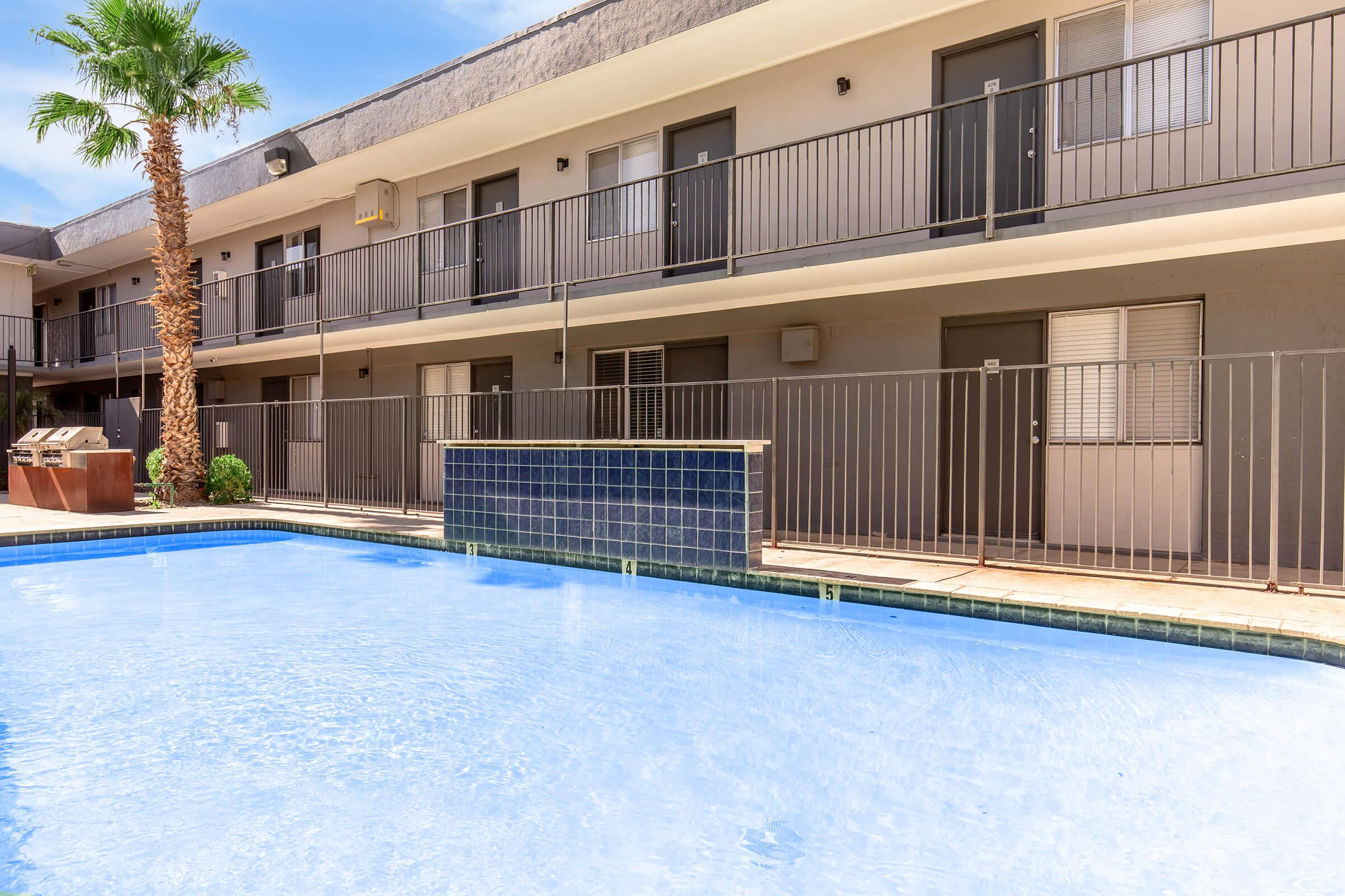 A view of a swimming pool in a courtyard surrounded by apartment buildings. The pool features blue tiles and is enclosed by a fence. In the background, several doors of the apartments are visible, along with a palm tree adding to the tropical ambiance.