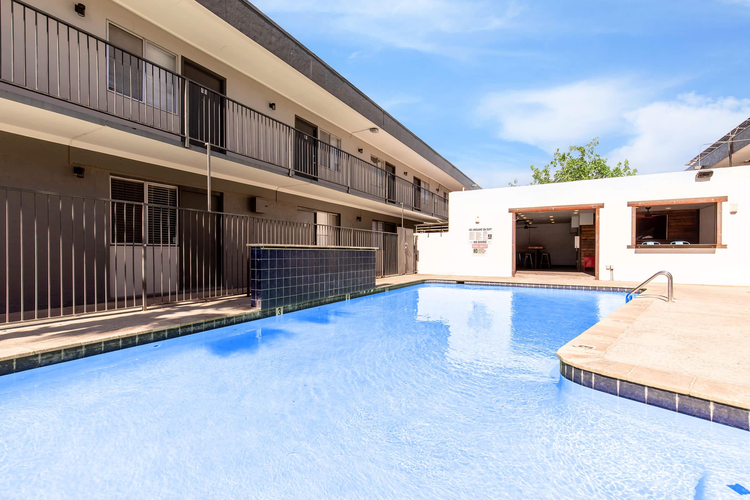 A view of a swimming pool in a courtyard surrounded by two-story residential buildings. The pool features clear blue water, and there are lounge areas visible. One building showcases sliding glass doors and balconies, while a communal area with seating is situated nearby, under a partly cloudy sky.