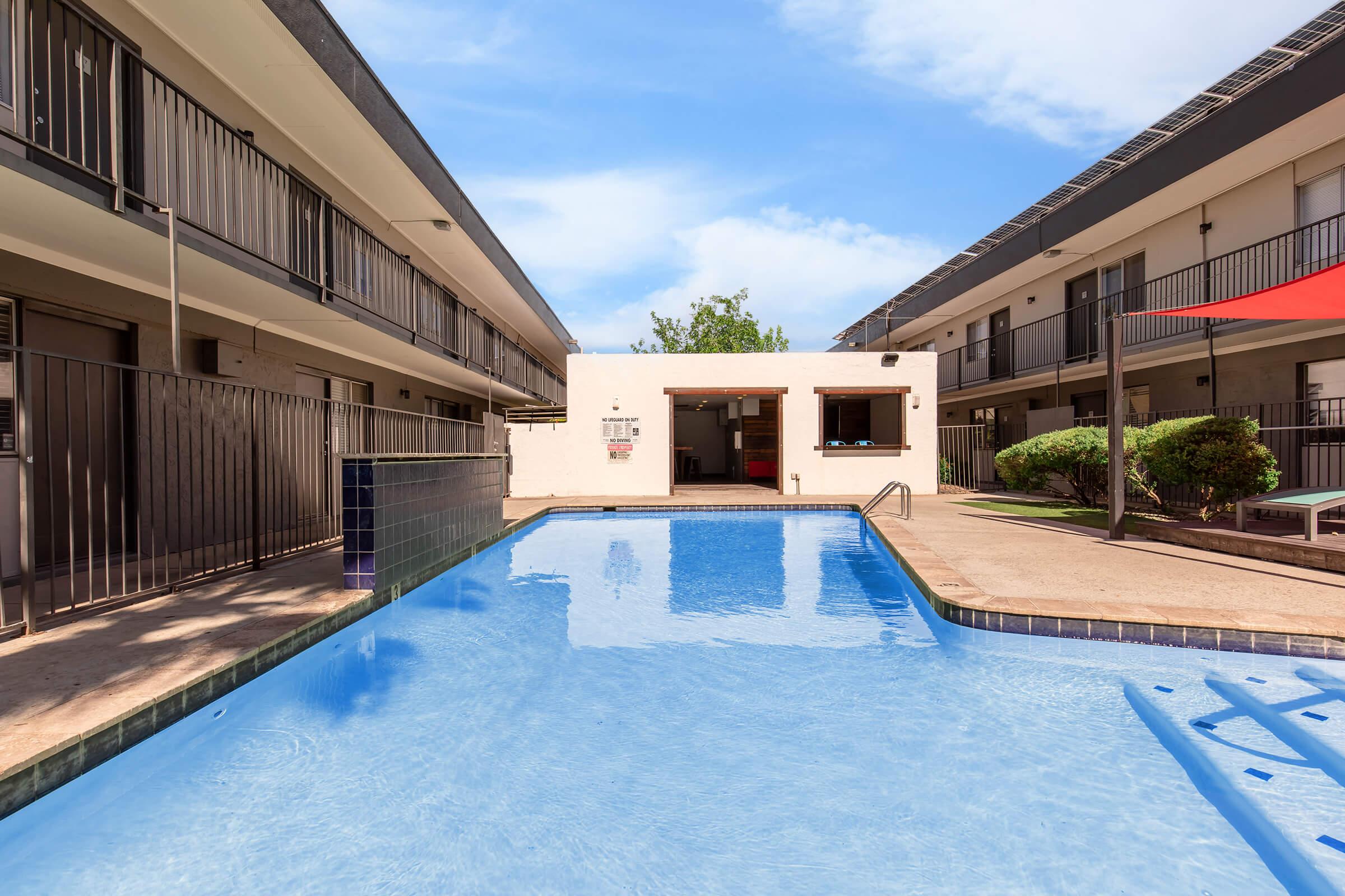 A swimming pool situated in the center of a courtyard surrounded by two-story apartment buildings. The pool features clear blue water and a shallow end with a ladder. Lush greenery and lounge chairs are visible nearby, and there’s a shaded area with a red canopy. The sky is bright with scattered clouds.