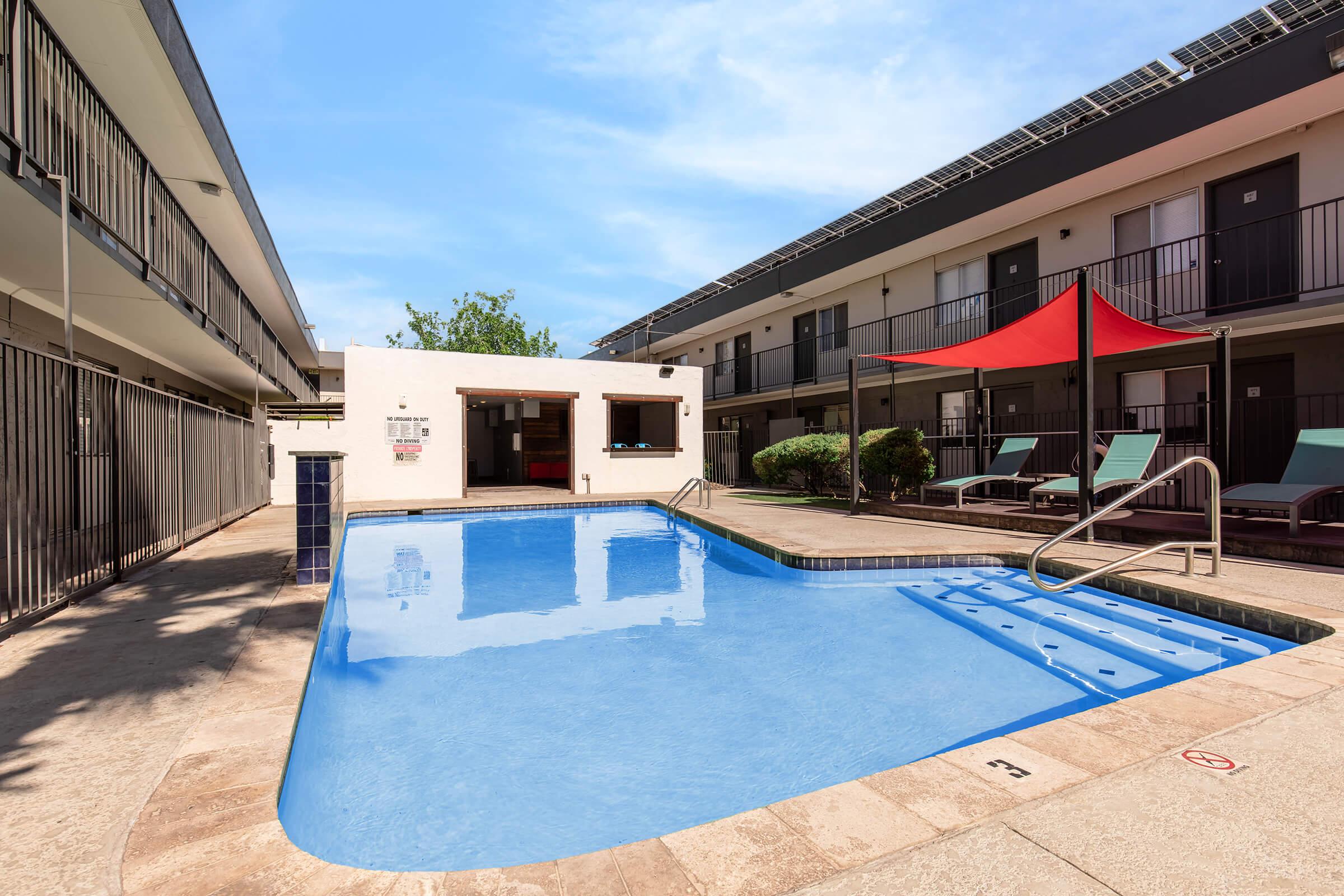 A clean outdoor pool area surrounded by two-story buildings. The pool features a shallow entry and a diving board. Lounge chairs line the poolside, and there is a shaded seating area with a red canopy. Solar panels are visible on the rooftops, and the sky is clear and blue.