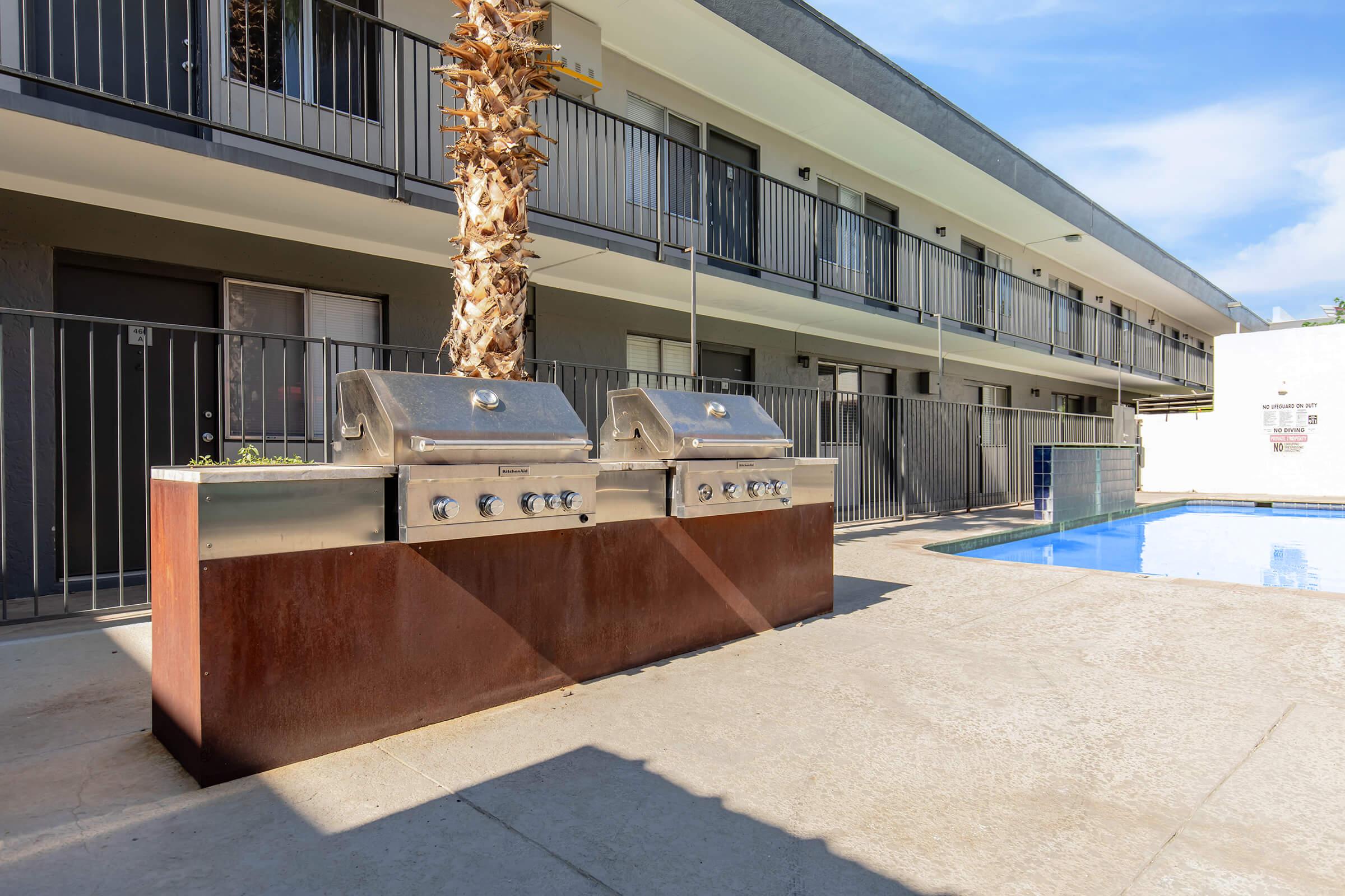 Outdoor grilling area featuring two stainless steel gas grills on a rust-colored stone platform, adjacent to a swimming pool. In the background, a two-story building with balconies and palm trees is visible, set against a clear blue sky.