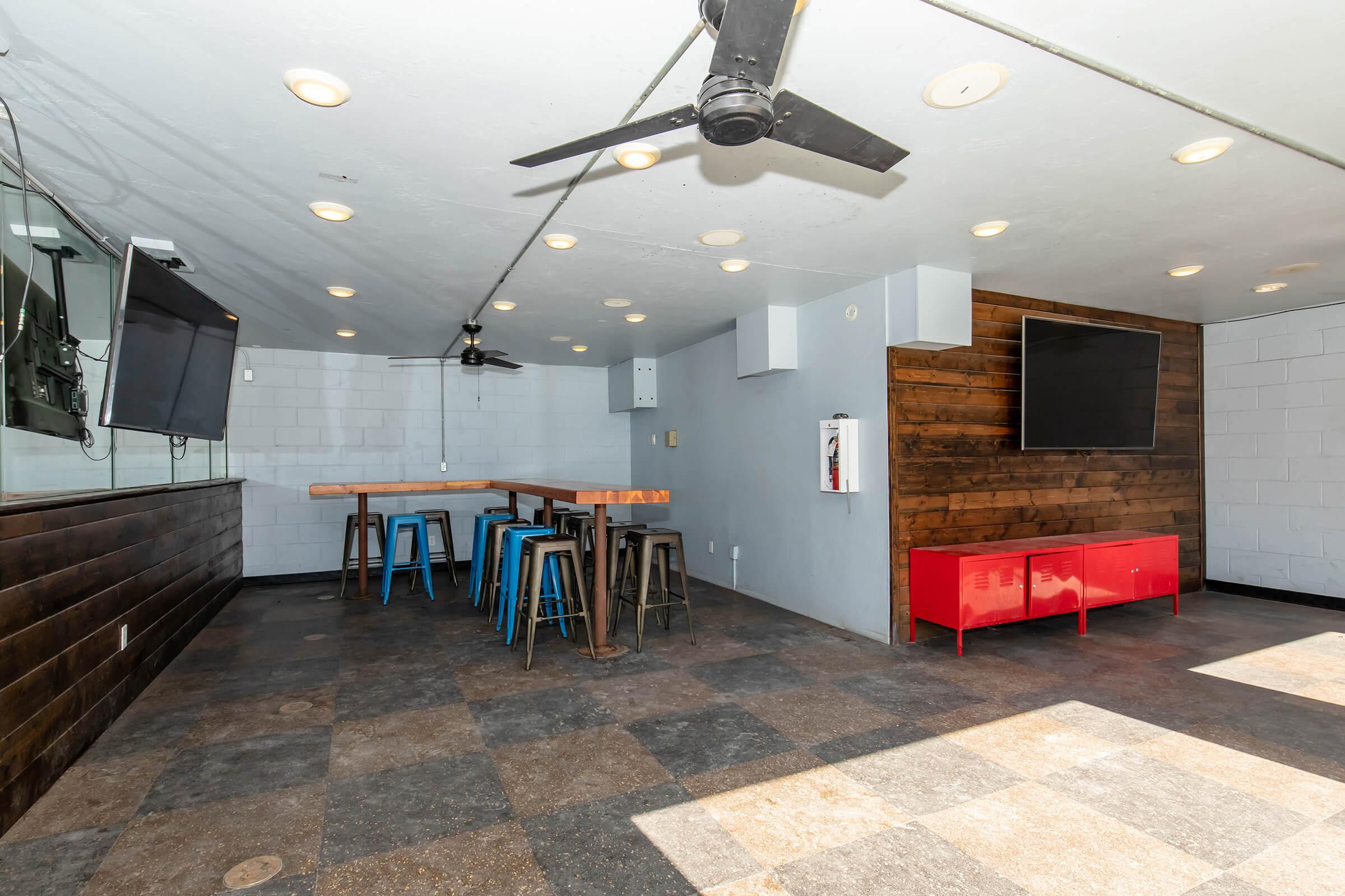 An empty, modern interior space featuring a dark tiled floor, wooden walls, and bright overhead lights. There are two flat-screen televisions mounted on the walls, a red storage unit, and a high table with metal stools. The area is well-lit and has a ceiling fan for airflow.