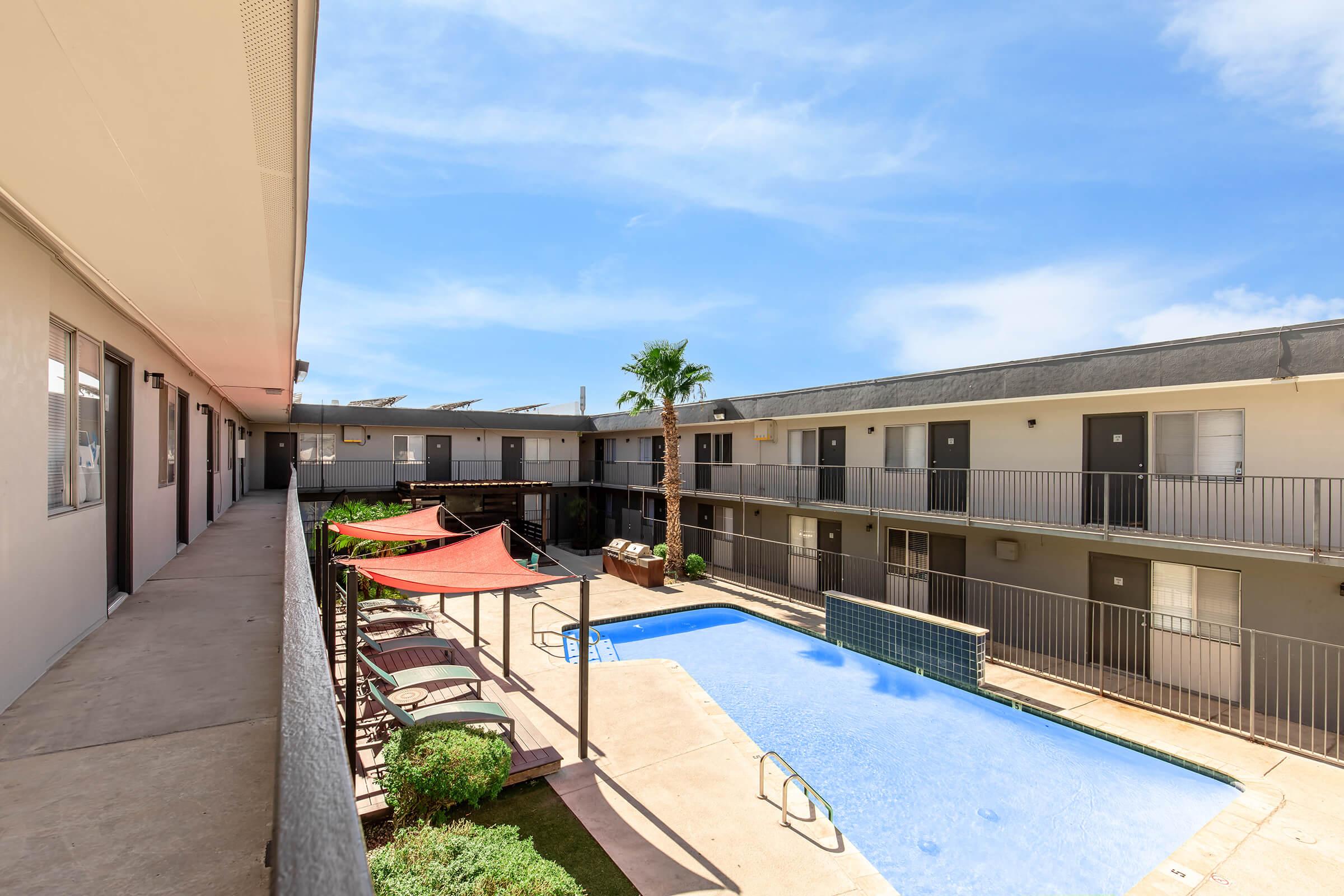 A view of a courtyard with a swimming pool surrounded by two-story apartment buildings. The pool area features lounge chairs and shaded seating under colorful sun umbrellas. Lush green landscaping adds to the inviting atmosphere under a clear blue sky.