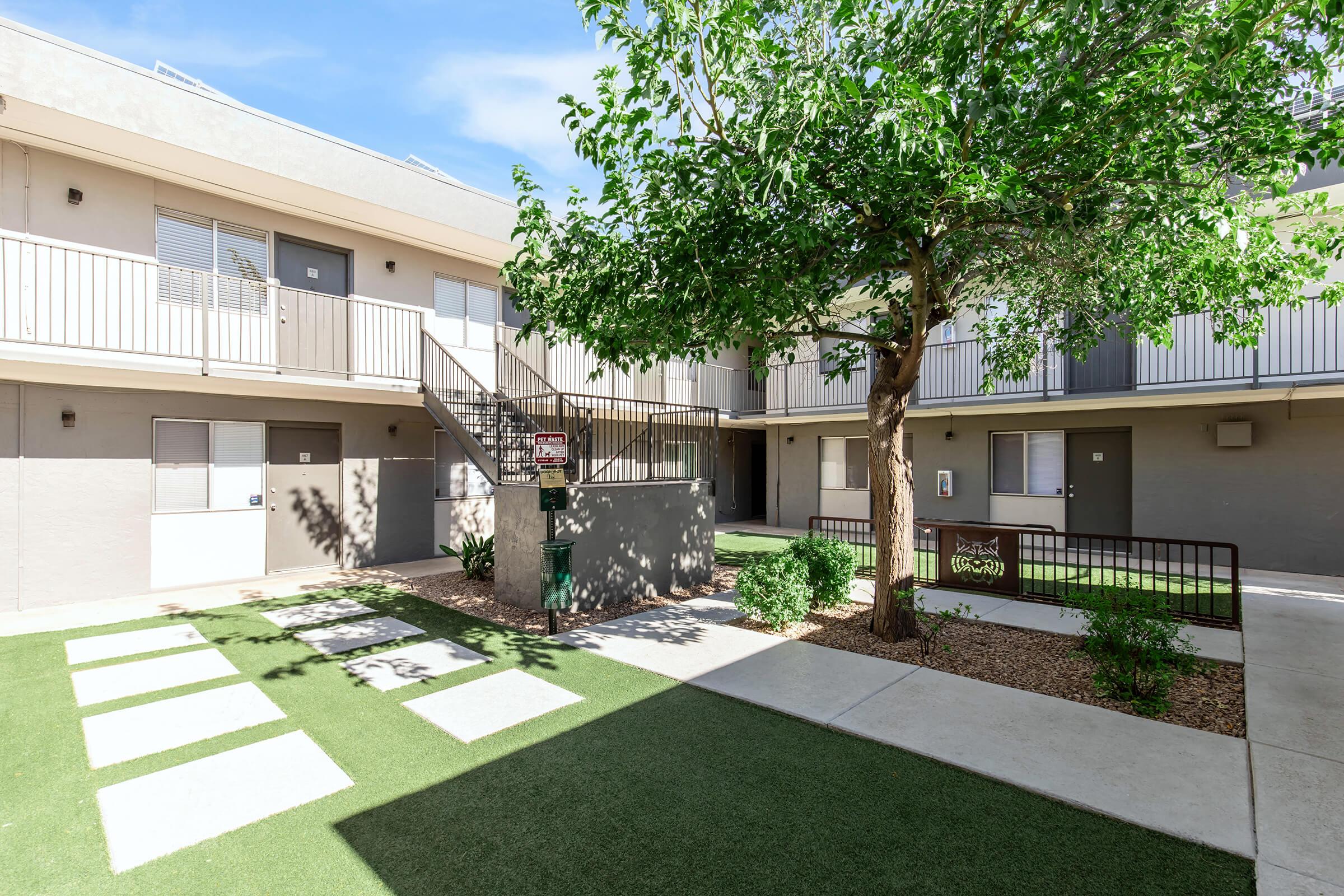 View of a courtyard in an apartment complex featuring a well-maintained grassy area, a small tree, and concrete walkways leading to the building entrances. There are multiple balconies visible, and the blue sky enhances the serene atmosphere of the community space.