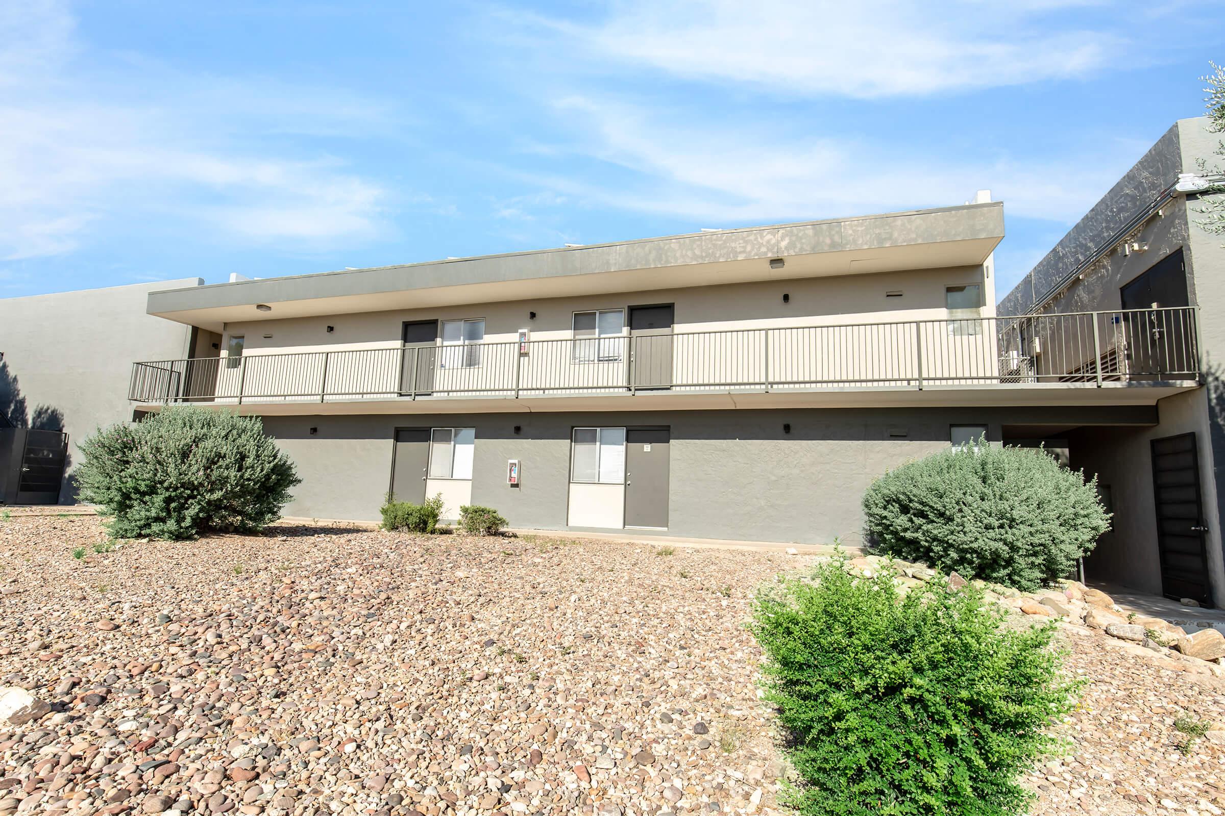 Two-story apartment building with a balcony on the second floor. The structure features a simple design with gray walls and multiple entrance doors. Surrounding the building are small shrubs and gravel landscaping under a clear blue sky.