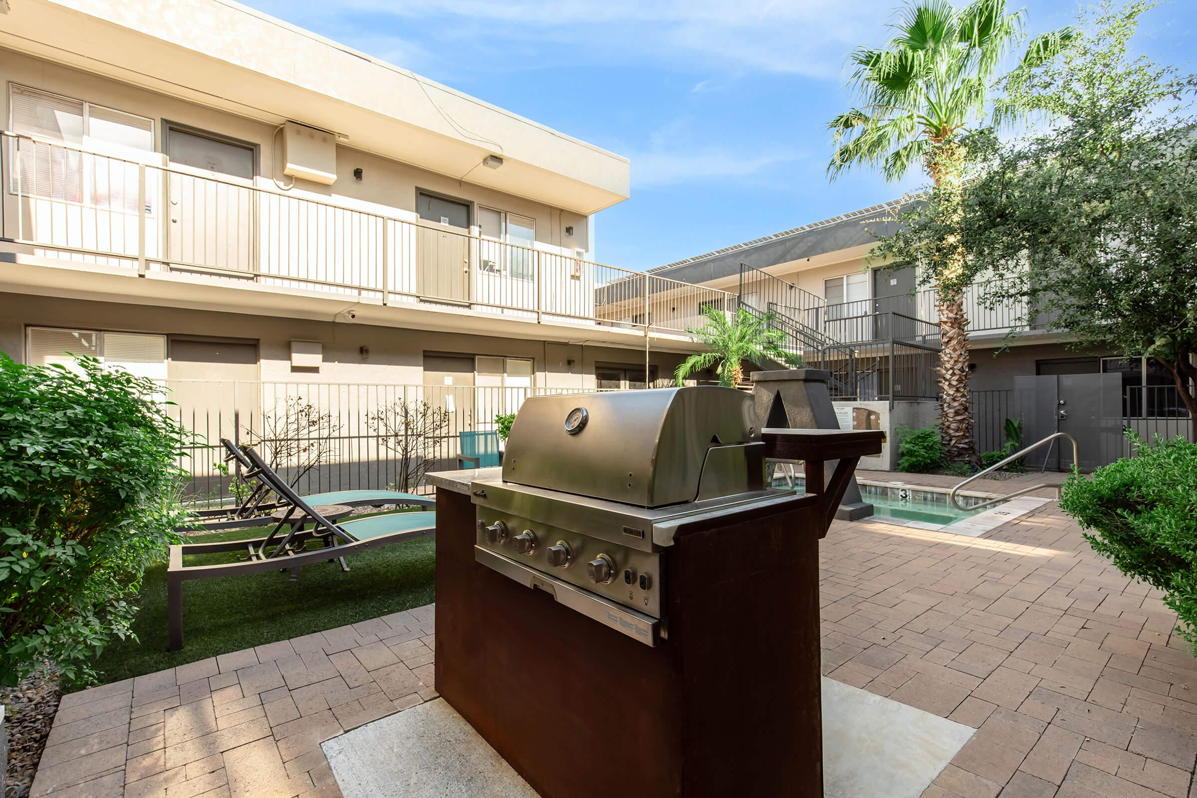 A barbecue grill sits in a landscaped courtyard area surrounded by low greenery, with an outdoor seating area and a swimming pool in the background. The setting features two-story apartment buildings with balconies, palm trees, and clear blue skies.