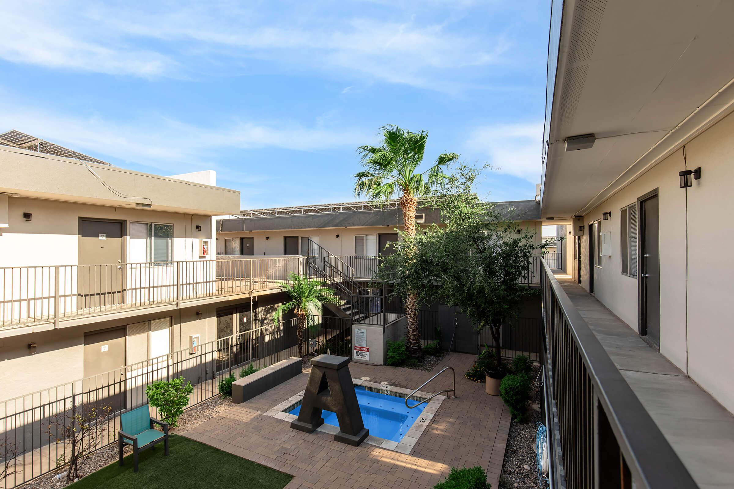 A mid-century style courtyard of an apartment complex featuring a small swimming pool, palm trees, and a landscaped area with grass and seating. The buildings are two stories, with balconies overlooking the courtyard, and a clear blue sky overhead.