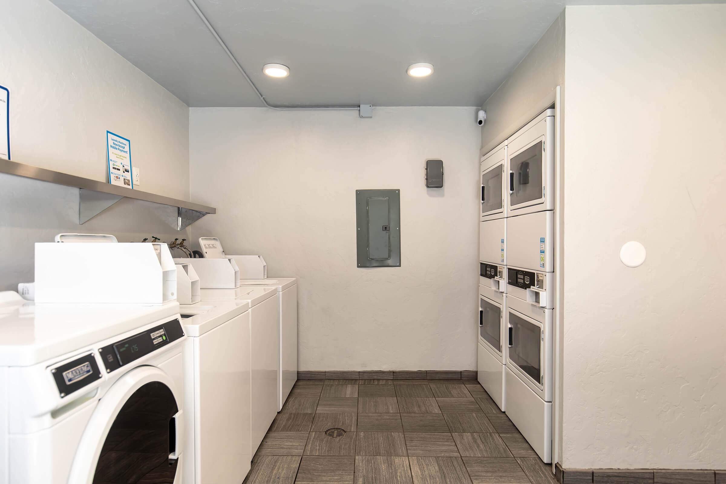 A clean laundry room featuring several white washing machines and dryers lined up against a light-colored wall. The space has a modern design with recessed lighting, a countertop for folding clothes, and a utility panel mounted on the wall. The floor is covered with square tiles.