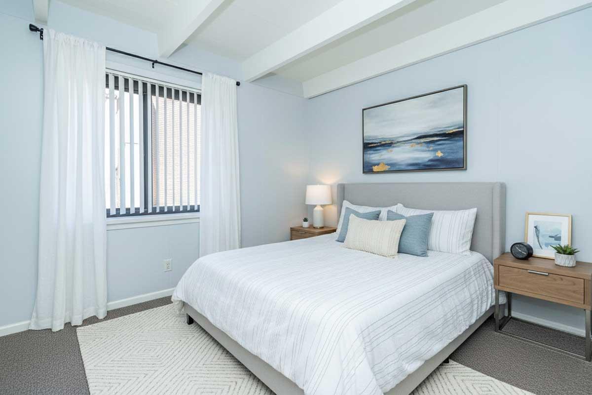 A cozy bedroom featuring a light blue color scheme, a neatly made bed with white and blue bedding, and a bedside table with a lamp and clock. Large windows with white curtains and vertical blinds allow natural light. A piece of abstract art hangs on the wall, adding a touch of color to the space.