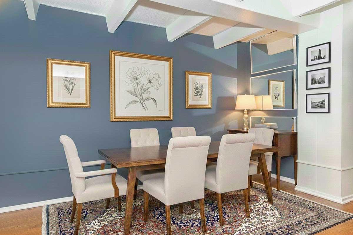 A beautifully arranged dining room featuring a wooden table surrounded by upholstered chairs. The walls are painted a soft blue, adorned with framed botanical prints. A stylish lamp illuminates the space, and a patterned rug adds warmth, complemented by a small side table and framed photographs on the adjacent wall.