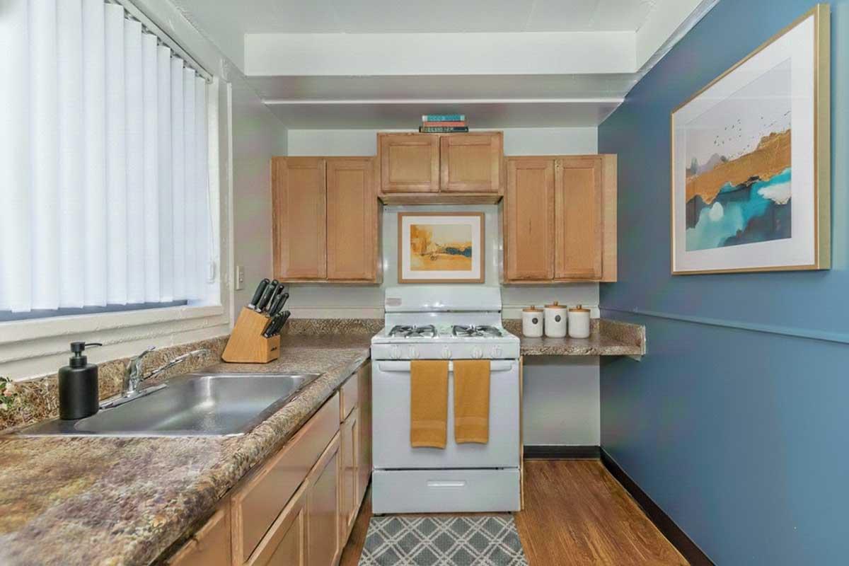 A modern kitchen featuring wooden cabinets, a stainless steel sink, and a gas stove with an oven. The countertop is made of brown and gray marble. A framed art piece hangs on the wall, and three white canisters are neatly arranged on the counter. Natural light filters through white blinds.