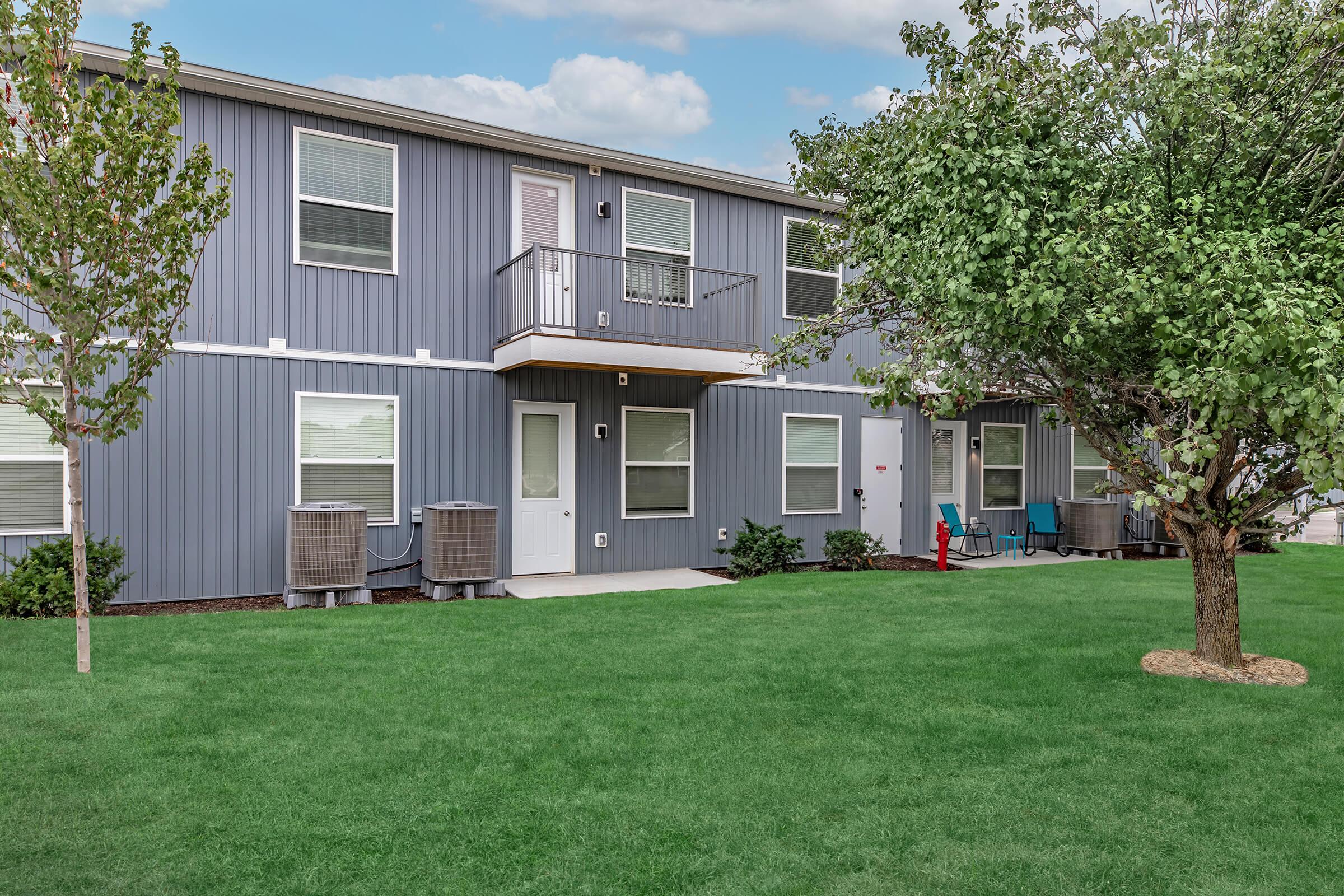 Two-story building with gray siding and white trim. The lower level has two doors and windows, with air conditioning units on either side. Lush green lawn in front, with a small tree and patio furniture visible. The sky is partly cloudy, creating a calm outdoor atmosphere.