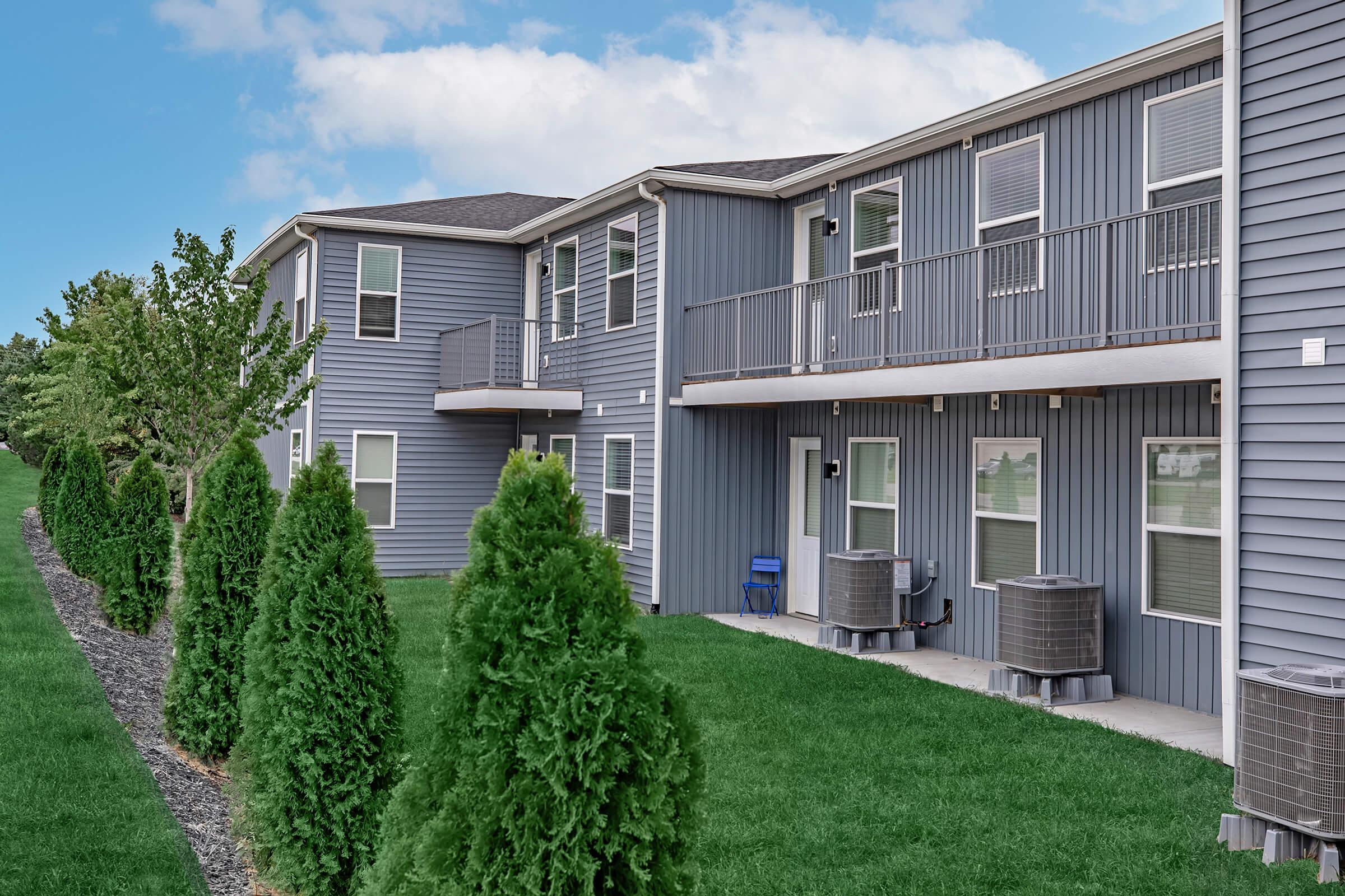 A row of modern, gray multi-family apartments with balconies, situated next to a well-maintained green lawn. Neatly trimmed evergreen shrubs line the walkway, and air conditioning units are placed outside each apartment. The sky is partly cloudy, giving a bright, inviting atmosphere.