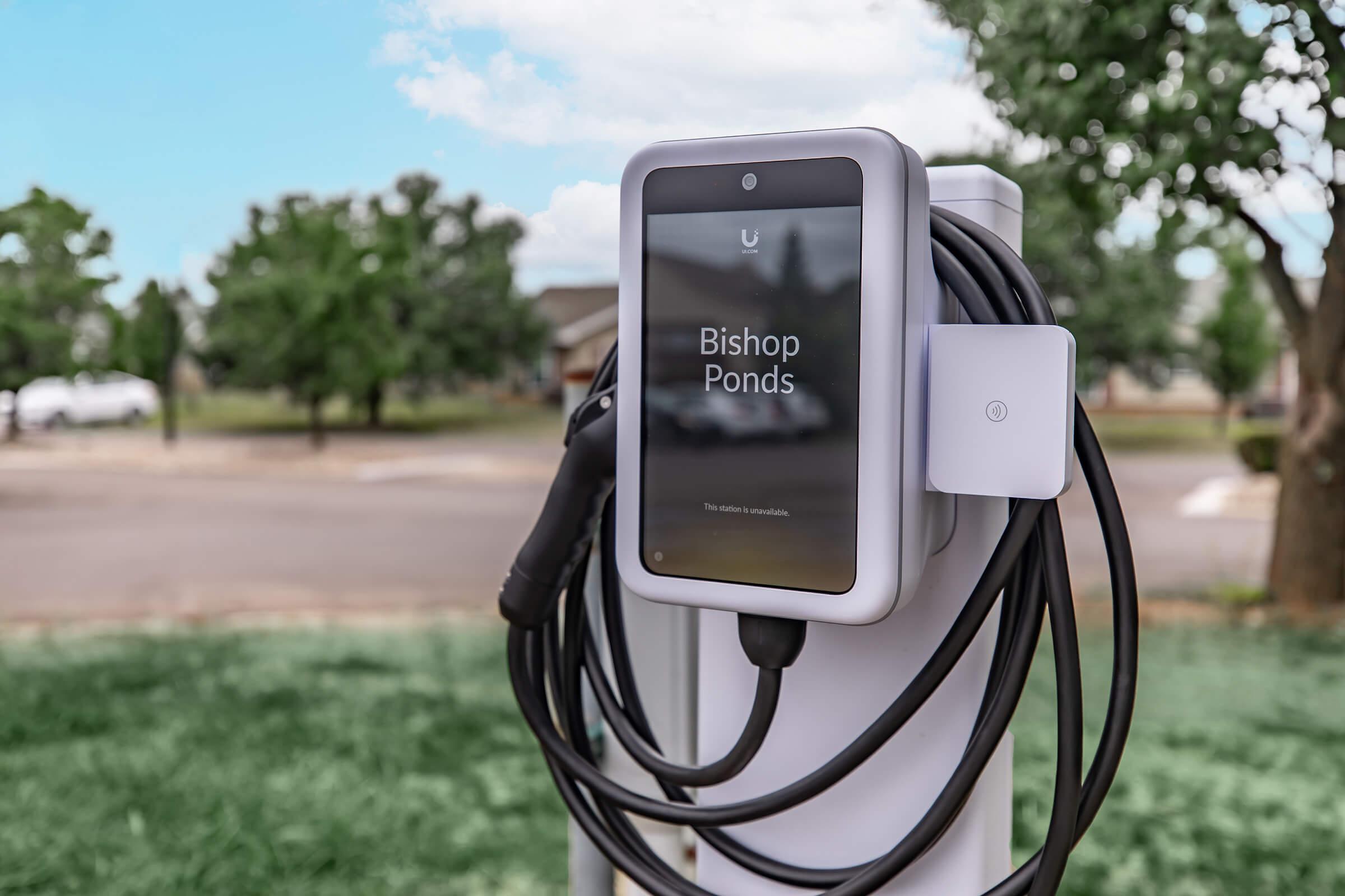 Electric vehicle charging station with a display screen showing "Bishop Ponds." The station has charging cables neatly coiled around it, set against a backdrop of a suburban area with trees and a cloudy sky.