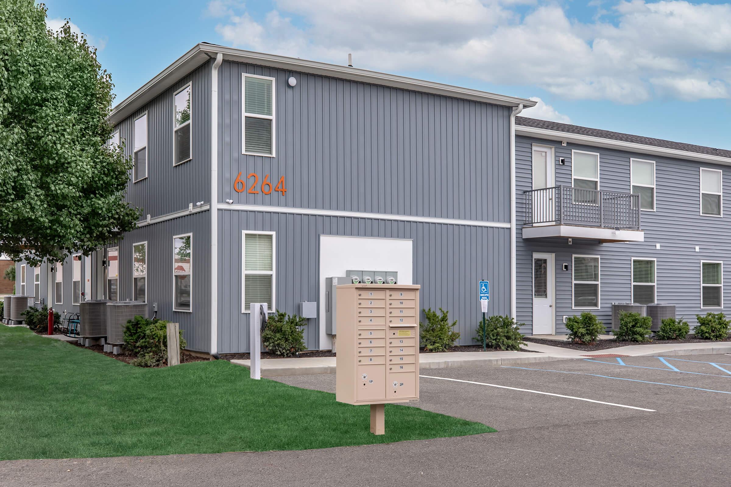 A modern two-story residential building with gray siding, featuring several windows and a small balcony. In front, a mailbox cluster stands on a grassy area, with the building number "6264" prominently displayed. The scene shows a parking lot with marked spaces and well-maintained landscaping.