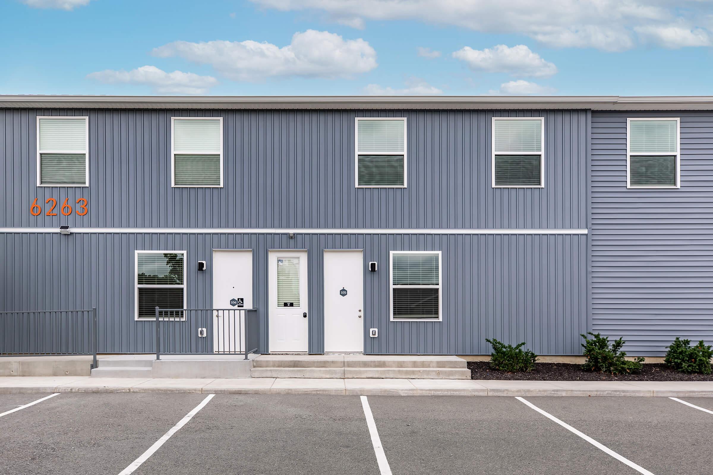 A modern two-story building with gray metal siding, featuring three windows on the upper floor and two white doors on the lower floor. The building is situated in a parking lot with clearly marked parking spaces, and the number '6263' is displayed prominently above the doors.