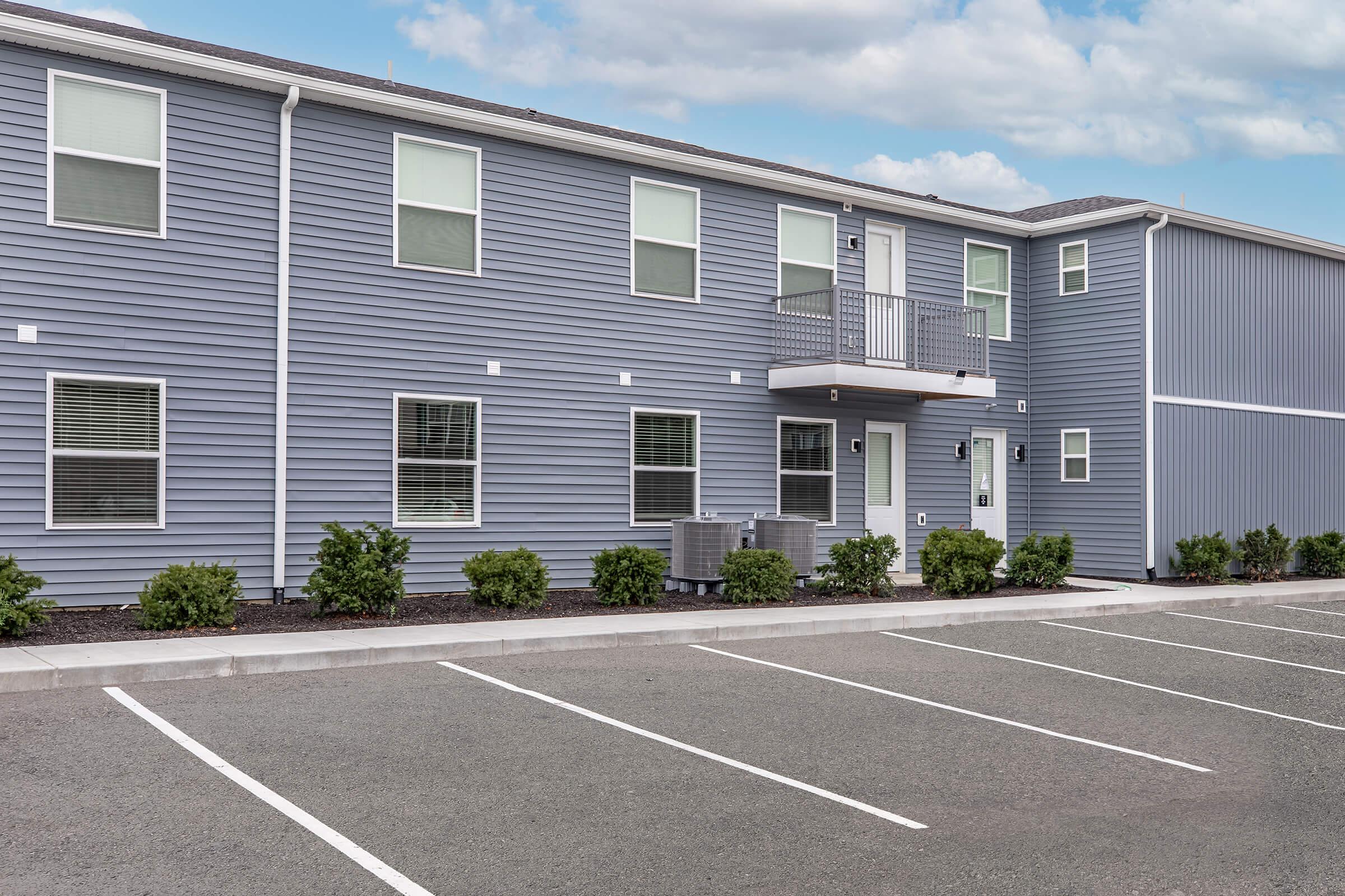 Two-story modern building with blue siding. The ground level features a small balcony above seating arrangements. Multiple windows are visible, along with a concrete parking area with several empty spaces. Small shrubs line the front of the building, and the sky is partly cloudy.