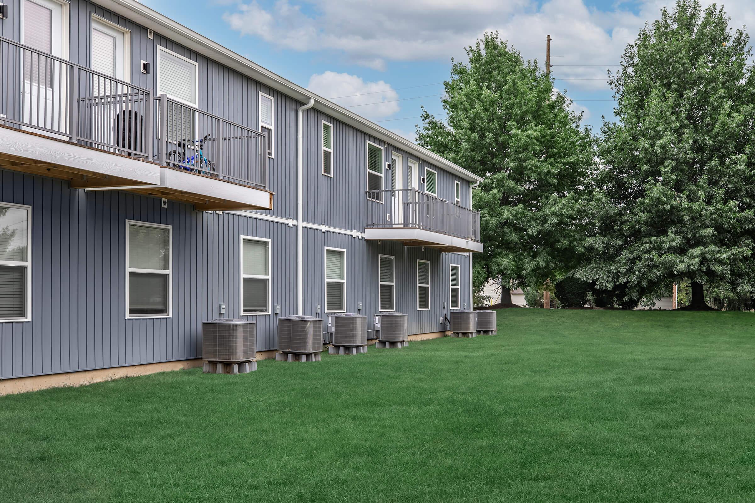 A two-story residential building with gray siding and balconies. Air conditioning units are placed along the ground level. A well-maintained green lawn is in front of the building, bordered by trees on one side under a partly cloudy sky.