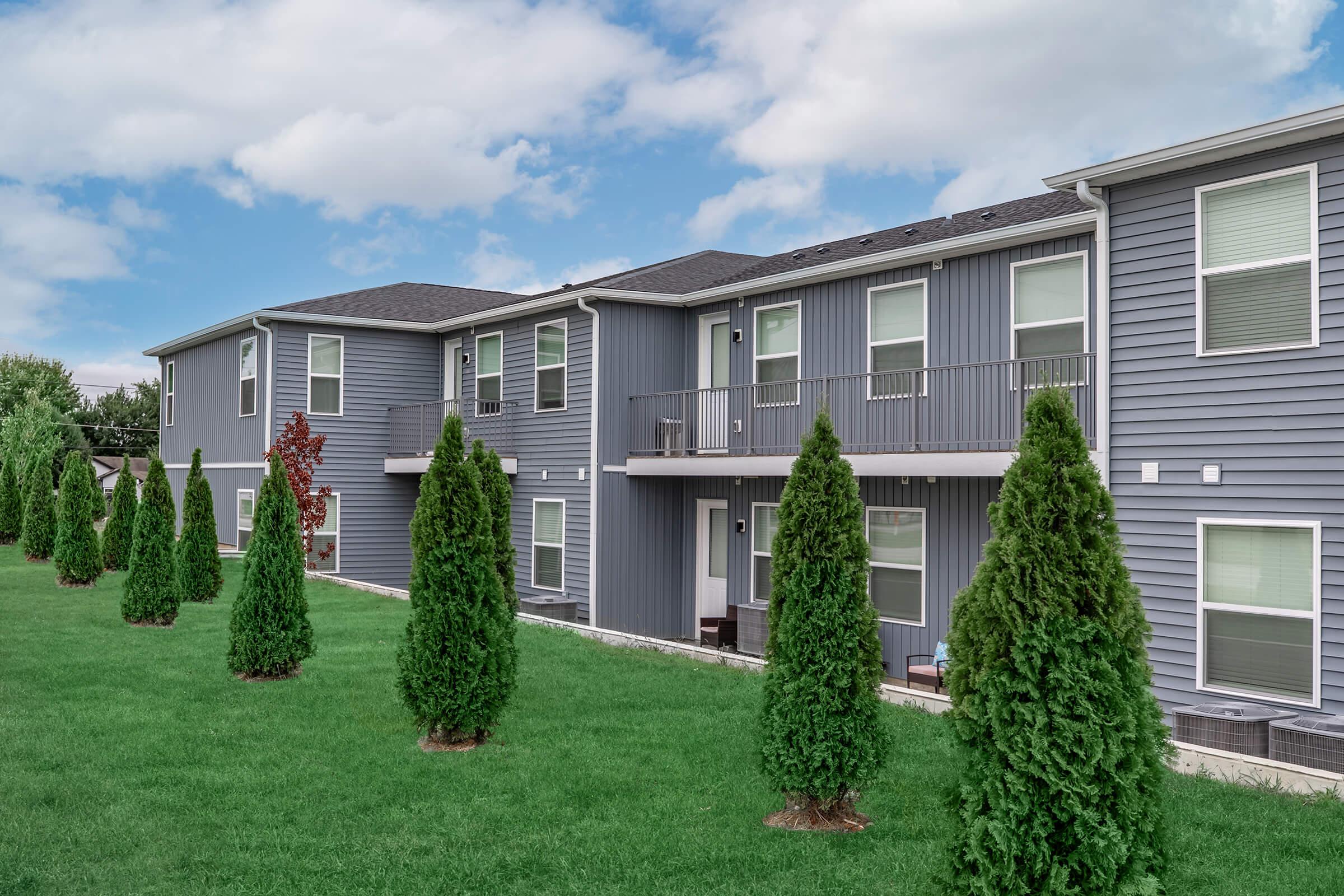 A row of modern, gray apartment buildings with multiple balconies, situated on a green lawn. Tall, narrow evergreen trees line the foreground, enhancing the outdoor aesthetic. The sky is partly cloudy, adding a serene atmosphere to the scene.