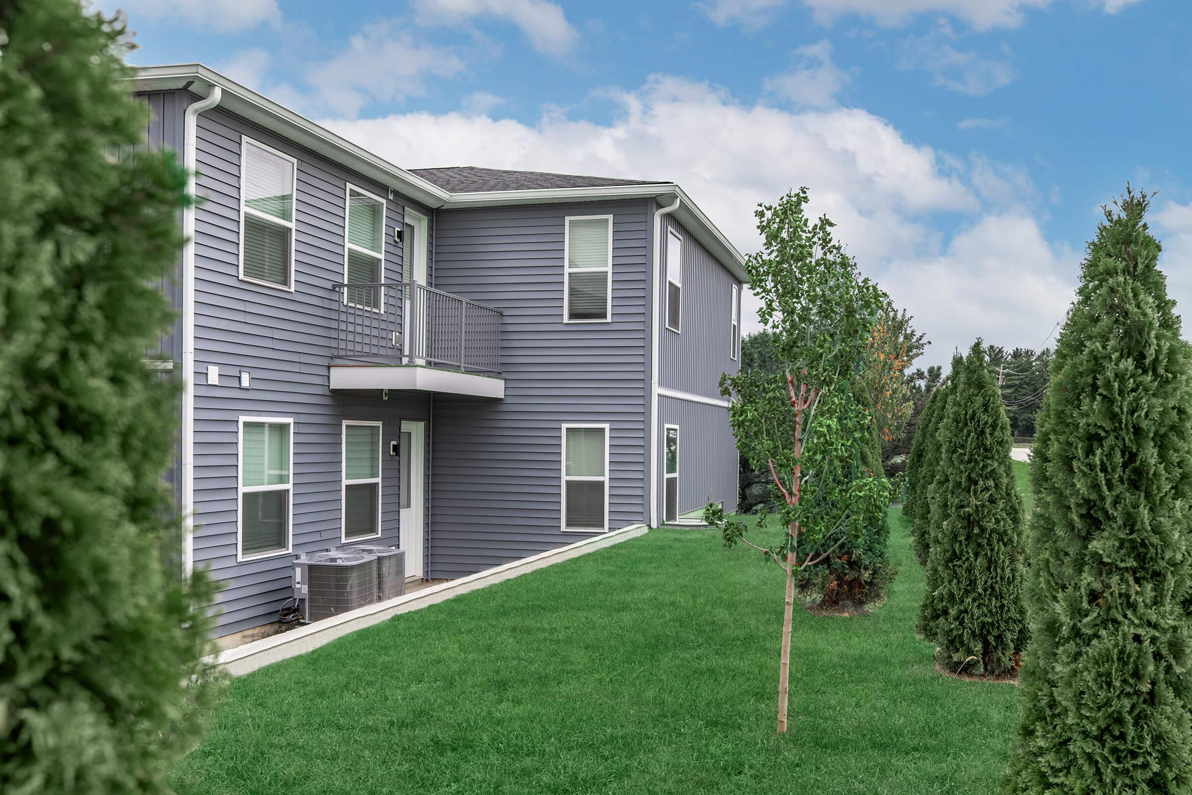 A modern two-story gray apartment building with a balcony, surrounded by well-maintained green grass and young trees. The sky is slightly overcast, creating a calm atmosphere. An air conditioning unit is visible next to the building, contributing to the contemporary design.