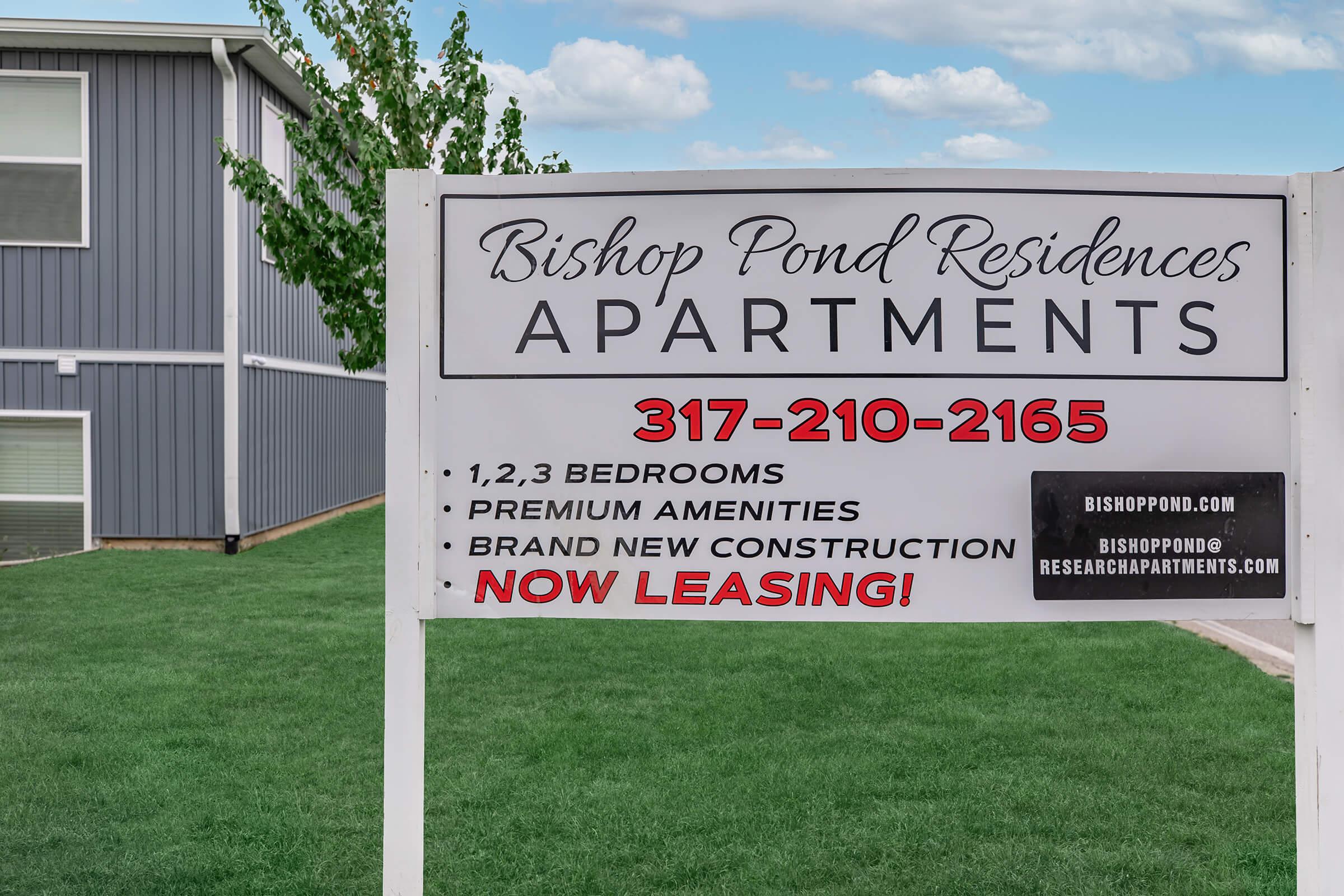Sign for Bishop Pond Residences Apartments advertising 1, 2, and 3-bedroom units with premium amenities. It mentions brand new construction and includes a phone number (317-210-2165) and website (bishoppond.com). The sign is placed in a grassy area near the apartment building.