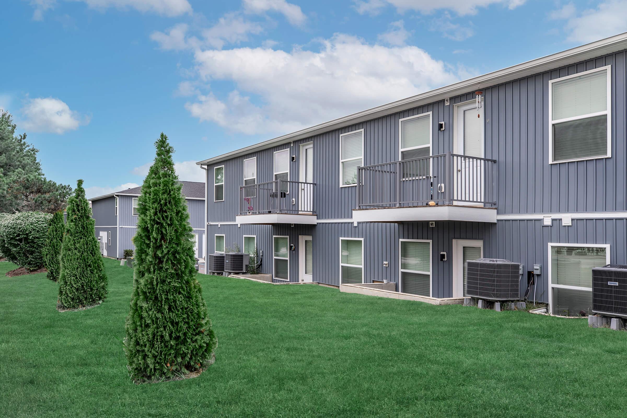 A modern apartment building featuring gray siding with two levels of balconies. Neatly trimmed evergreen trees line a lush green lawn in the foreground. Air conditioning units are visible on the ground level, and there are clouds in a blue sky above.