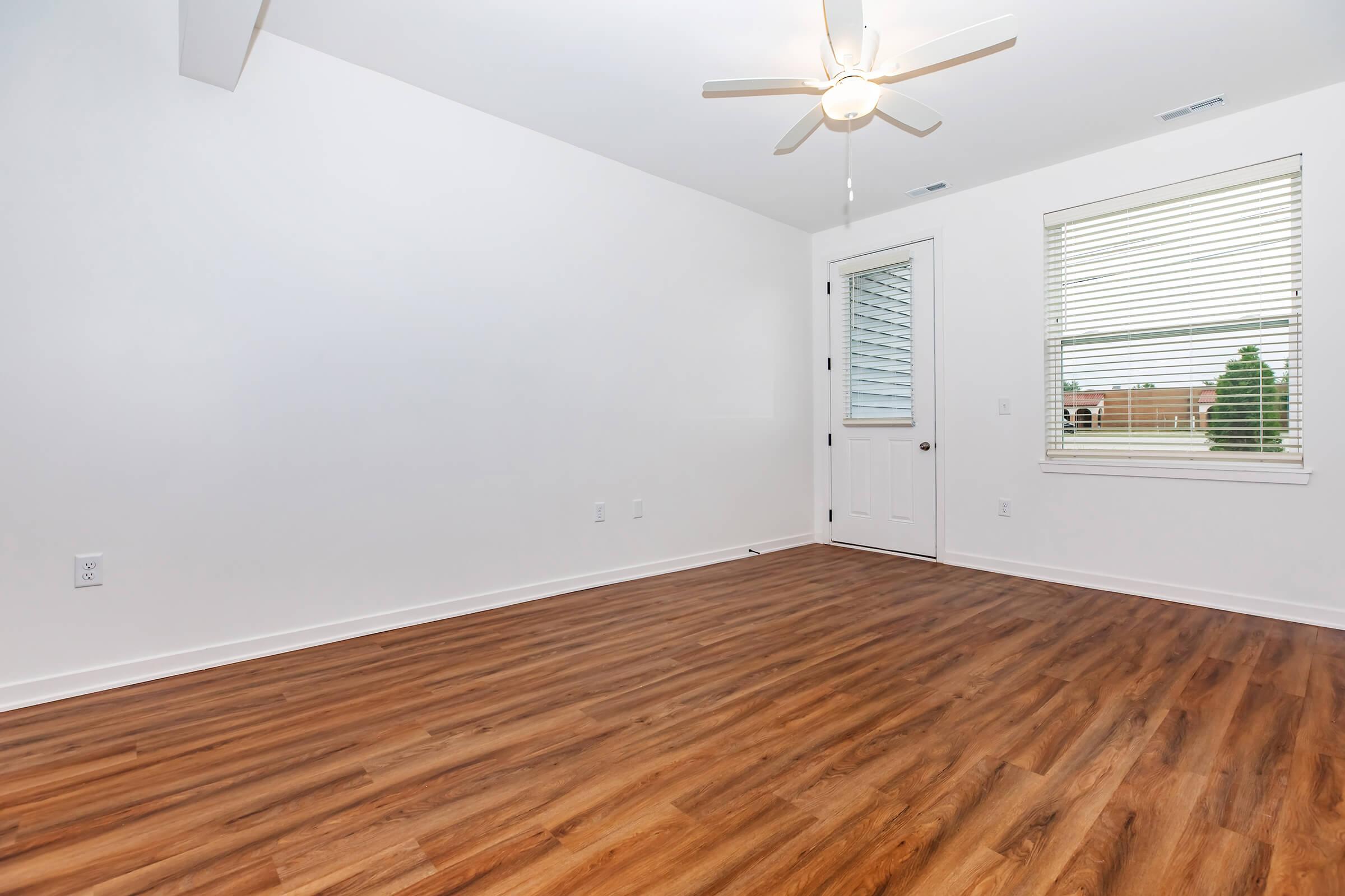 A bright, empty room with a light wood floor, a ceiling fan, and a window with blinds. The walls are painted white, and there's a door leading outside. The space appears modern and clean, suitable for various uses.