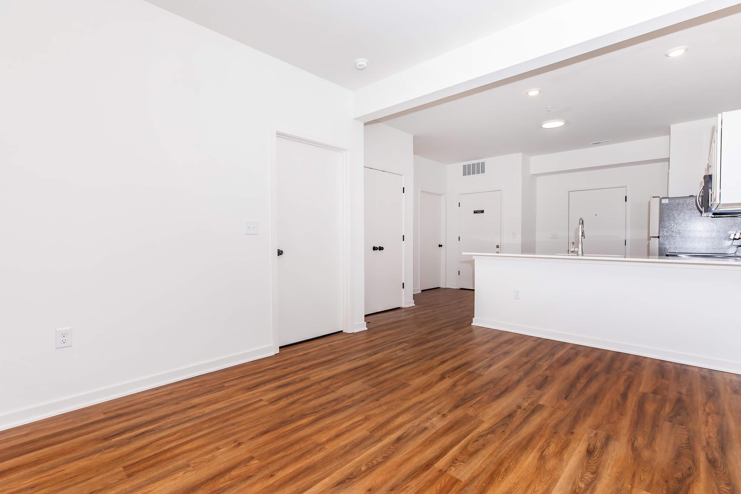 A spacious, empty apartment interior featuring light-colored walls, a wood-like floor, and a kitchen area with stainless steel appliances. There are multiple doors leading to other rooms, and the lighting is bright, creating an inviting atmosphere.