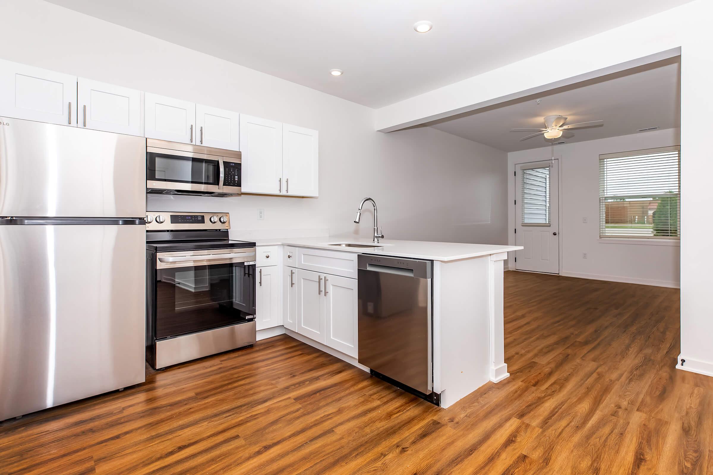Modern kitchen featuring stainless steel appliances including an oven, microwave, and dishwasher. The white cabinetry contrasts with the wooden floor. A spacious countertop with a sink is in the center, while an open area leads to a living space with large windows and a ceiling fan. Bright and inviting atmosphere.