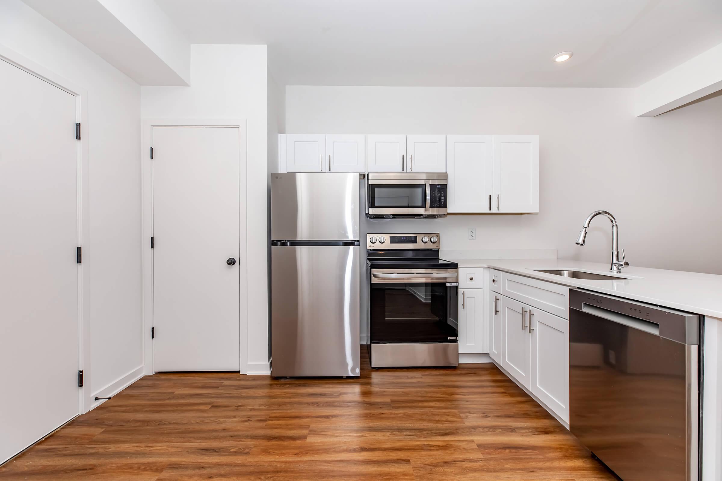 Modern kitchen featuring stainless steel appliances, including a refrigerator, oven, and dishwasher. The cabinetry is white, and there is a clean countertop with a sink. The floor is wooden, contributing to a bright and open atmosphere. Neutral colors dominate the space, enhancing its contemporary design.