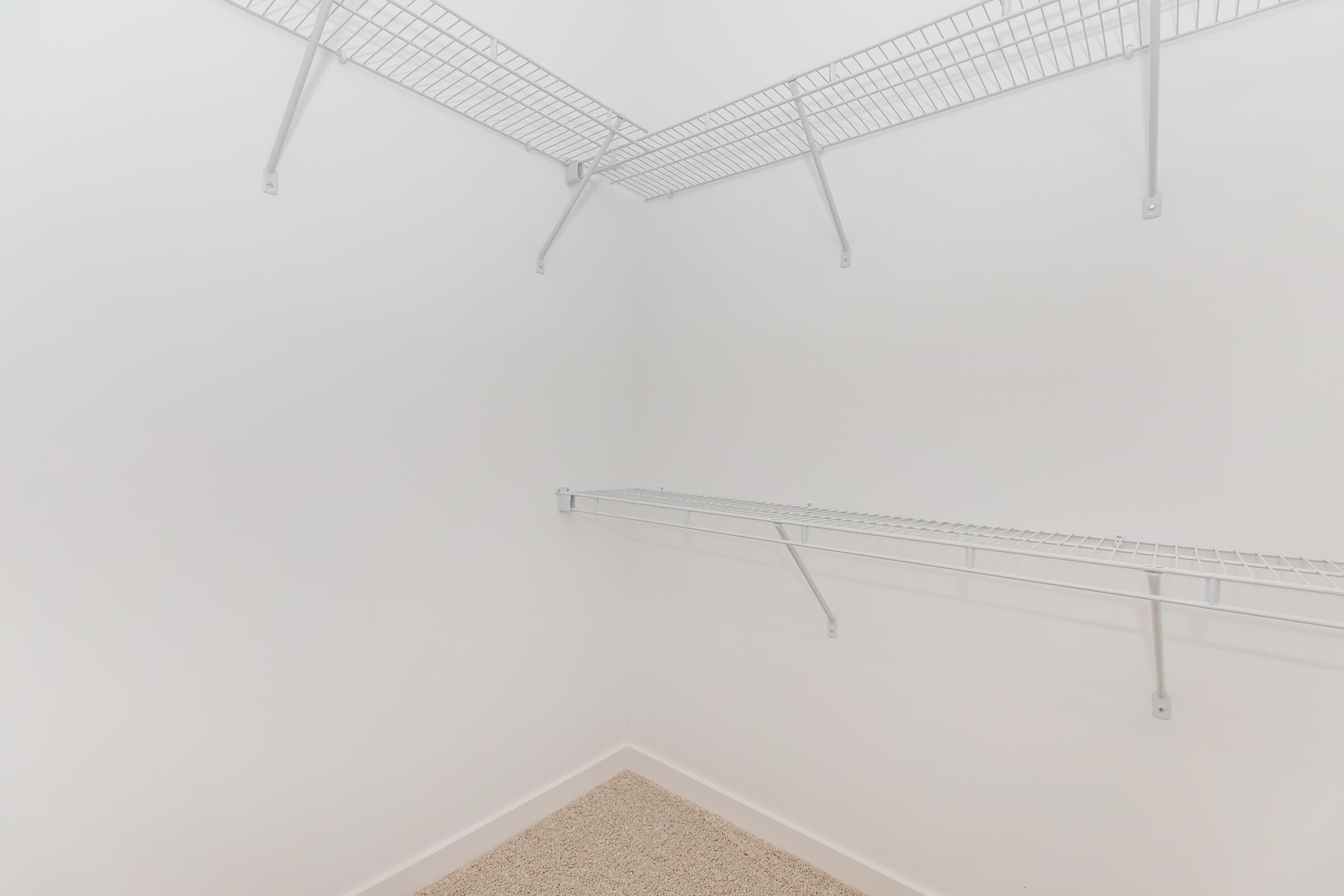 A clean, empty closet featuring white wire shelving mounted on the walls. The walls are bare and painted white, with a light-colored carpeted floor. The minimalistic design suggests ample space for storage or organization.