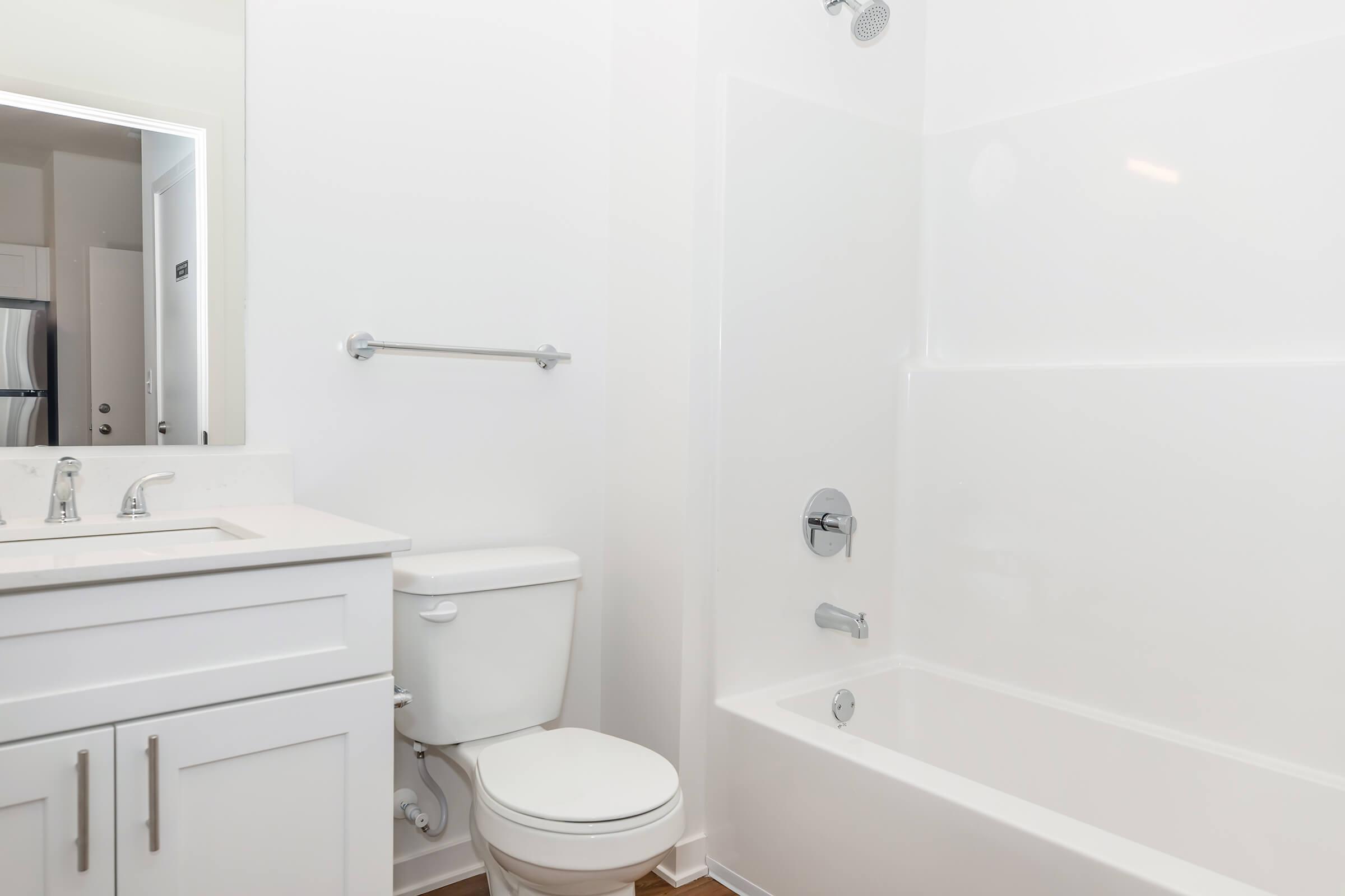 A clean and modern bathroom featuring a white bathtub, a toilet, a sink with a vanity, and a chrome faucet. The walls and bathtub are also white, creating a bright, minimalist look, with a large mirror above the sink and a towel bar on the wall.