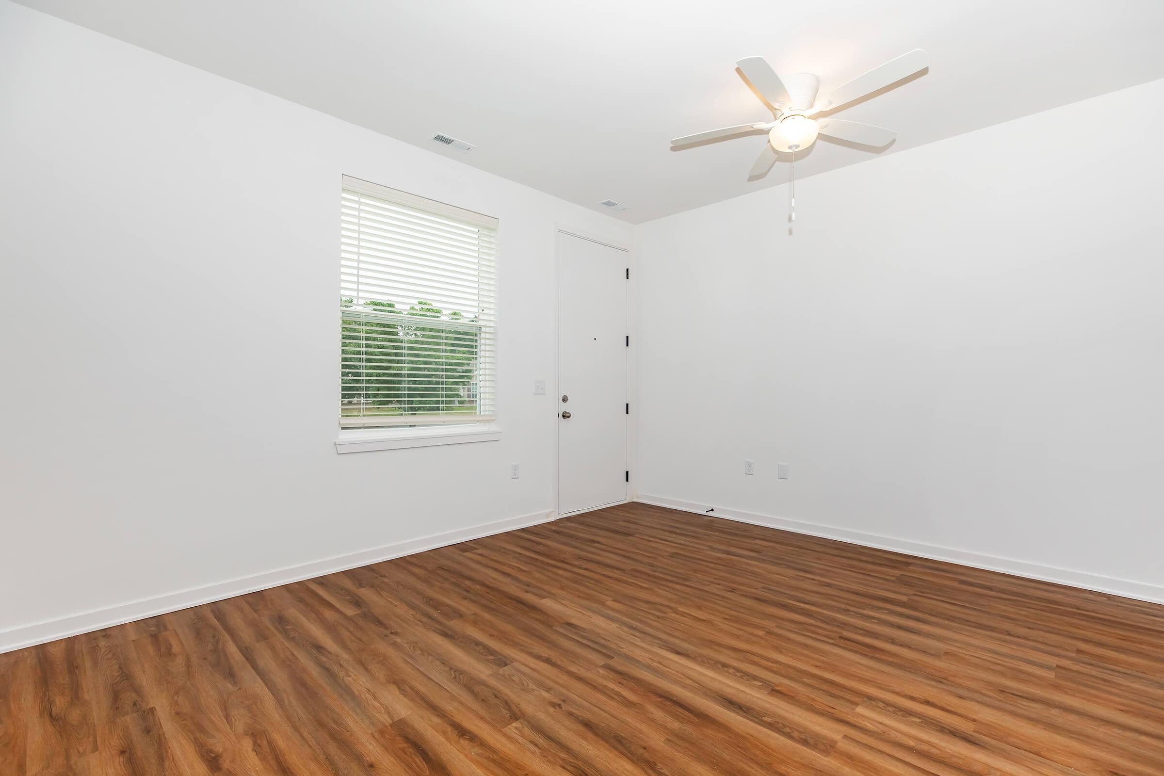 A bright, empty room featuring a wooden floor, white walls, and a ceiling fan. There is a window with blinds allowing natural light, and a door is visible on the right side of the image. The space appears clean and minimalist, suitable for various purposes.