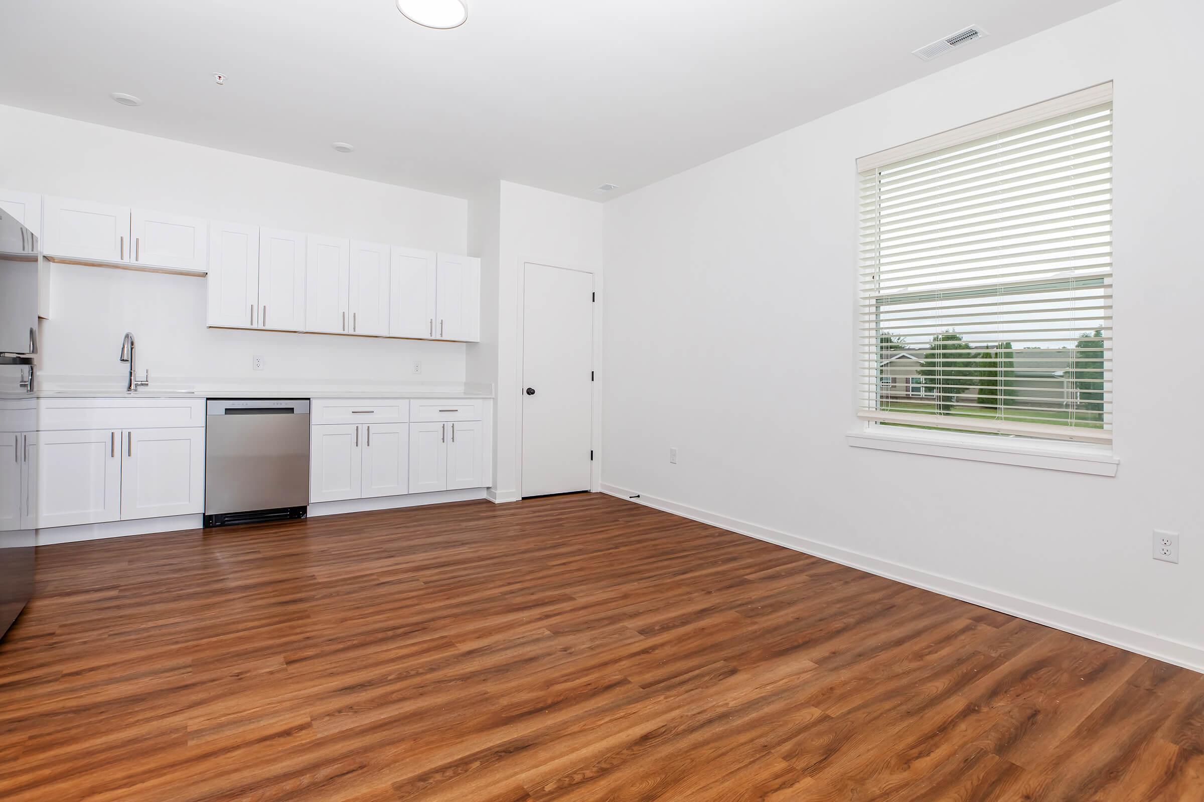 A modern kitchen and dining area featuring light wood floors, white cabinetry, a stainless steel refrigerator, and a dishwasher. The room has a large window with blinds, allowing natural light to flow in, and a plain white wall.