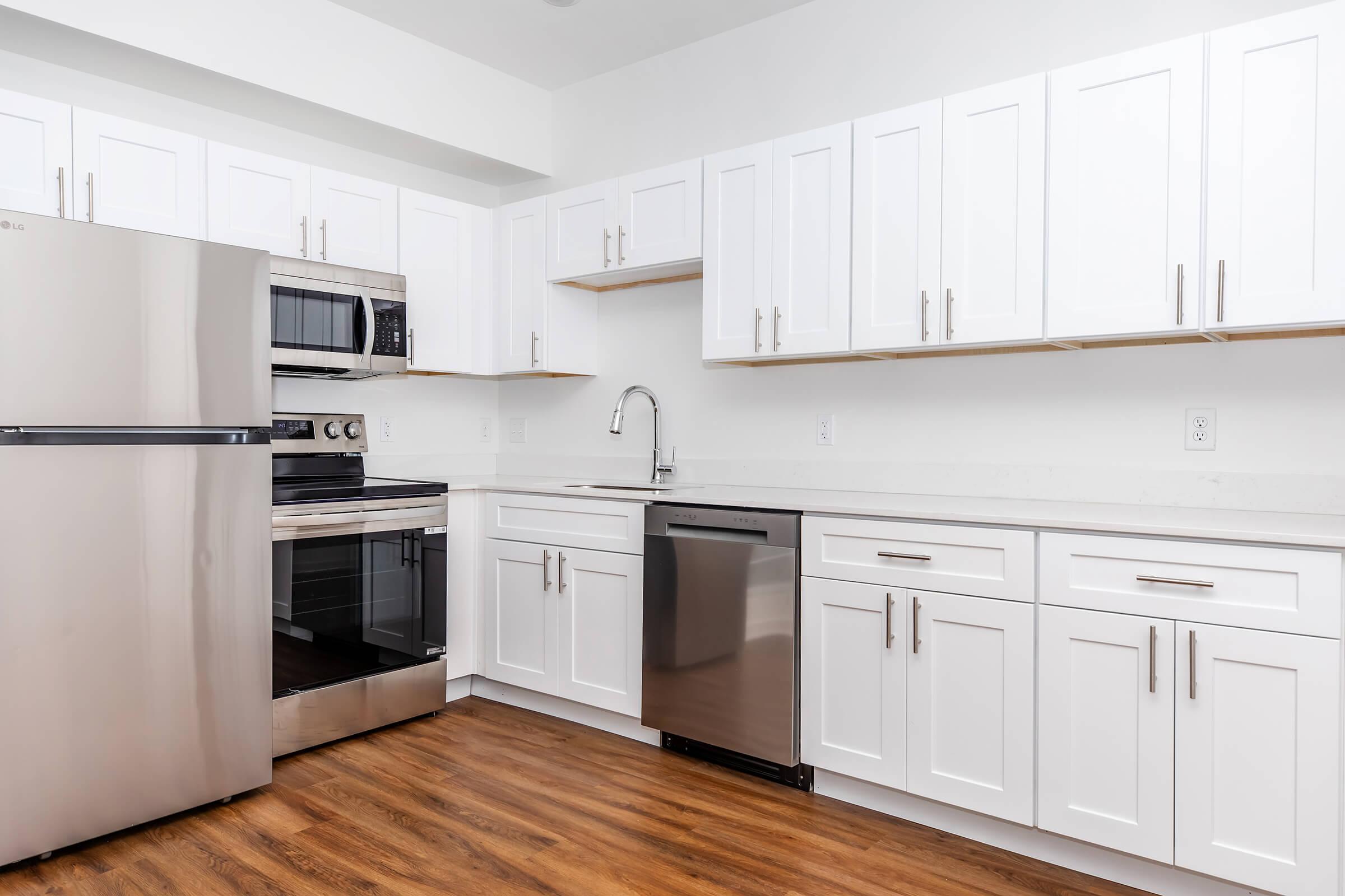 Modern kitchen featuring white cabinets, stainless steel appliances including a refrigerator, oven, and dishwasher, with a sink and minimalist design. The countertop is light-colored, and the floor has a wooden finish, creating a clean and contemporary look.