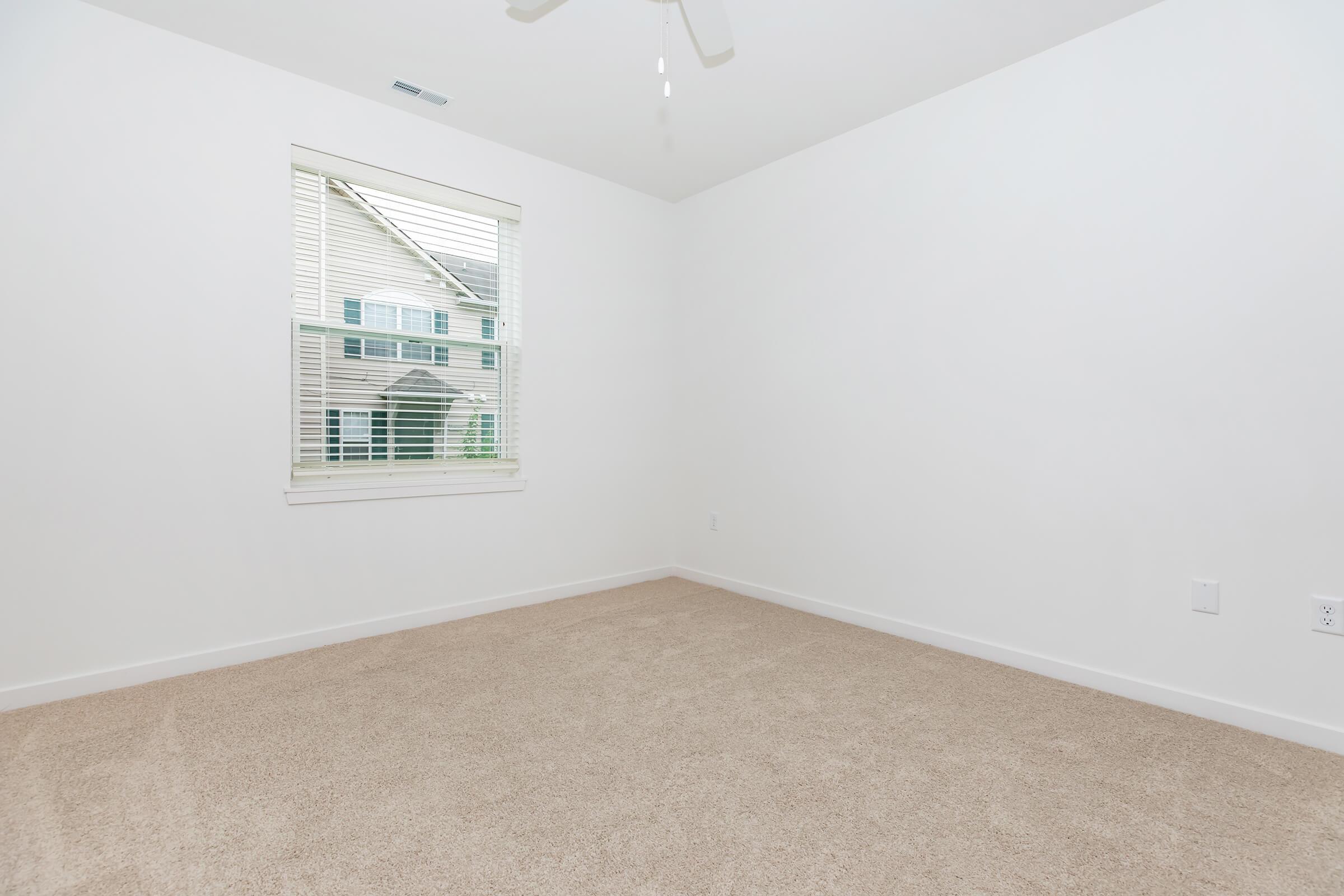 Empty room with light beige carpet, white walls, and a window with blinds. The view from the window shows a house exterior. A ceiling fan is visible, and the space has a clean, minimalist appearance.