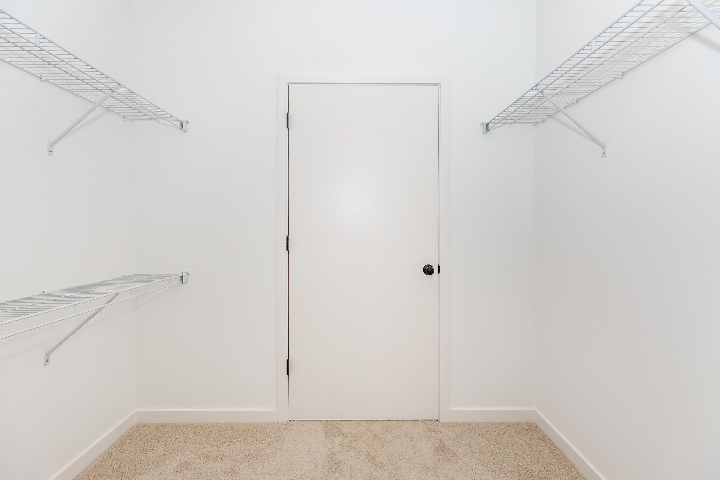 A vacant closet with white walls and a closed white door. Two metal wire shelves are mounted on each side of the closet, and the floor is covered in beige carpet. The space appears empty and uncluttered, creating a tidy and minimalist atmosphere.