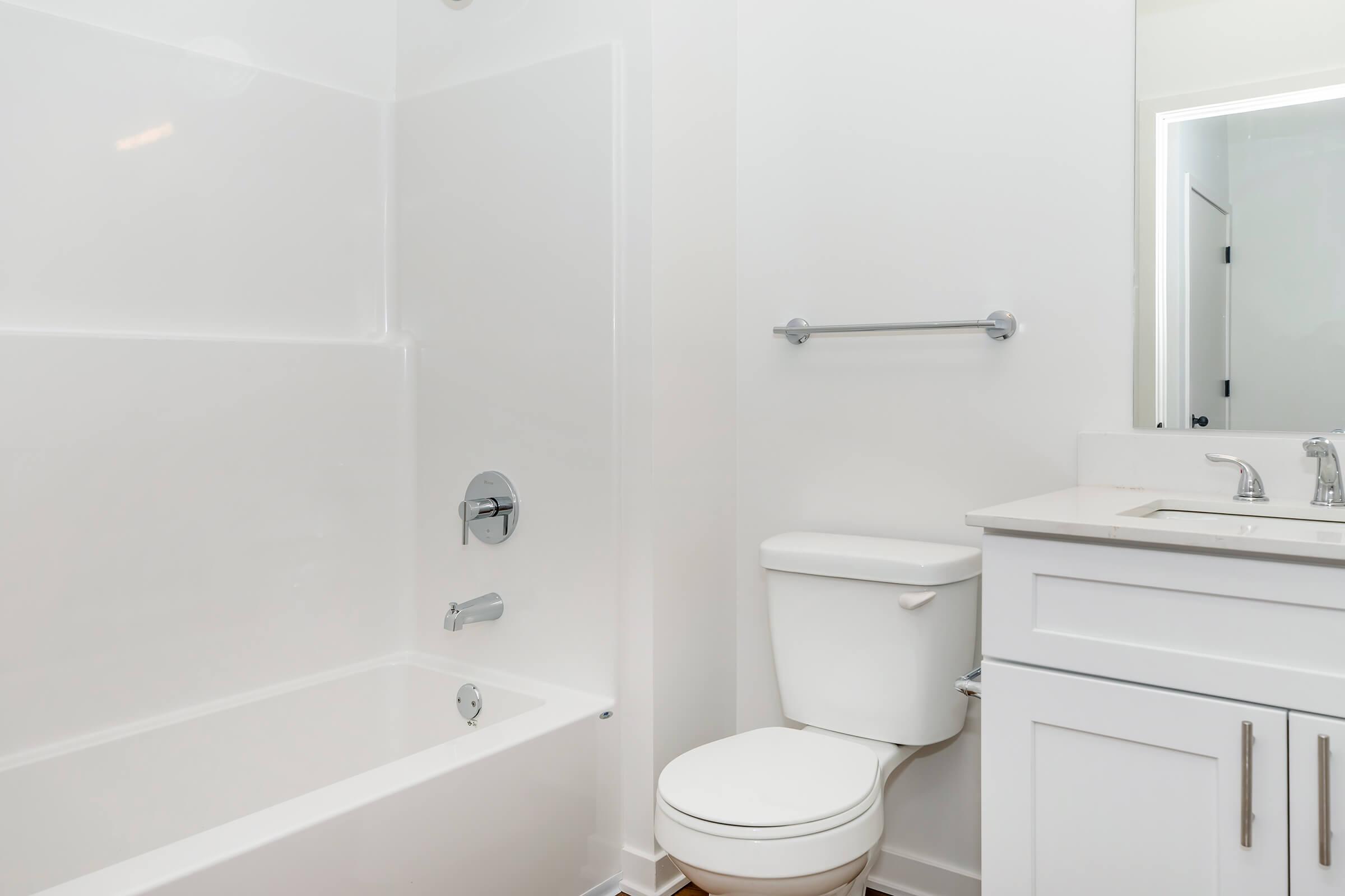 A clean and modern bathroom featuring a white bathtub with a shower combo, a toilet, and a sleek vanity with a sink. The walls and fixtures are bright and minimalistic, creating an inviting and spacious atmosphere. Light enters from a door leading to another area.