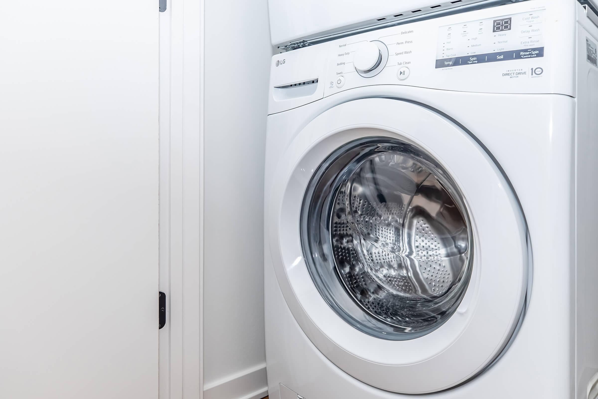 A modern white washing machine with a transparent front door, showcasing the drum inside, situated against a light-colored wall. The machine features digital controls and a sleek design, indicating a contemporary laundry space.