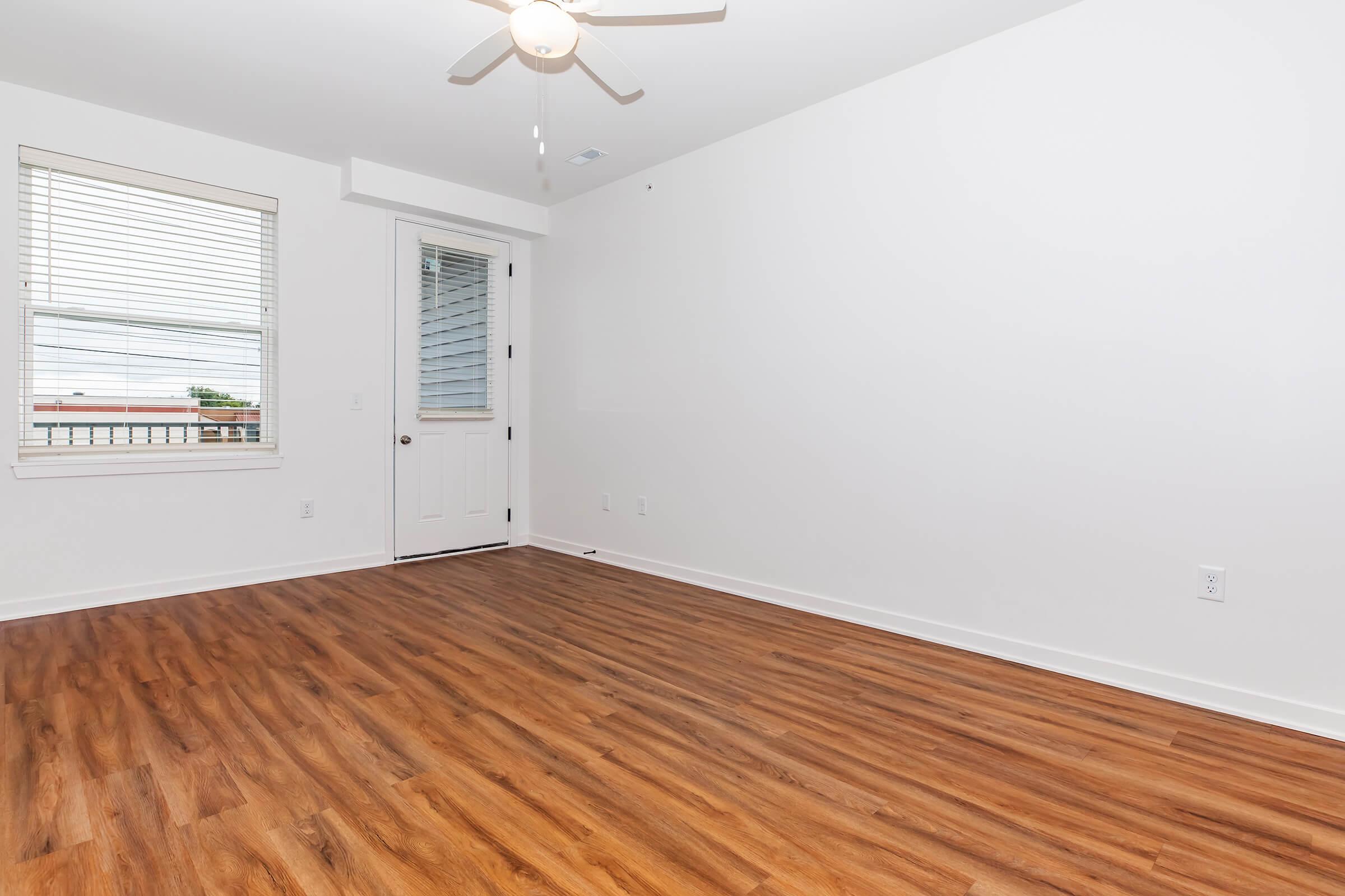 A spacious, empty room with light wood flooring, a ceiling fan, and a single window with blinds. The walls are painted white, and there is a door leading to another area. Natural light fills the space, creating a bright and inviting atmosphere.
