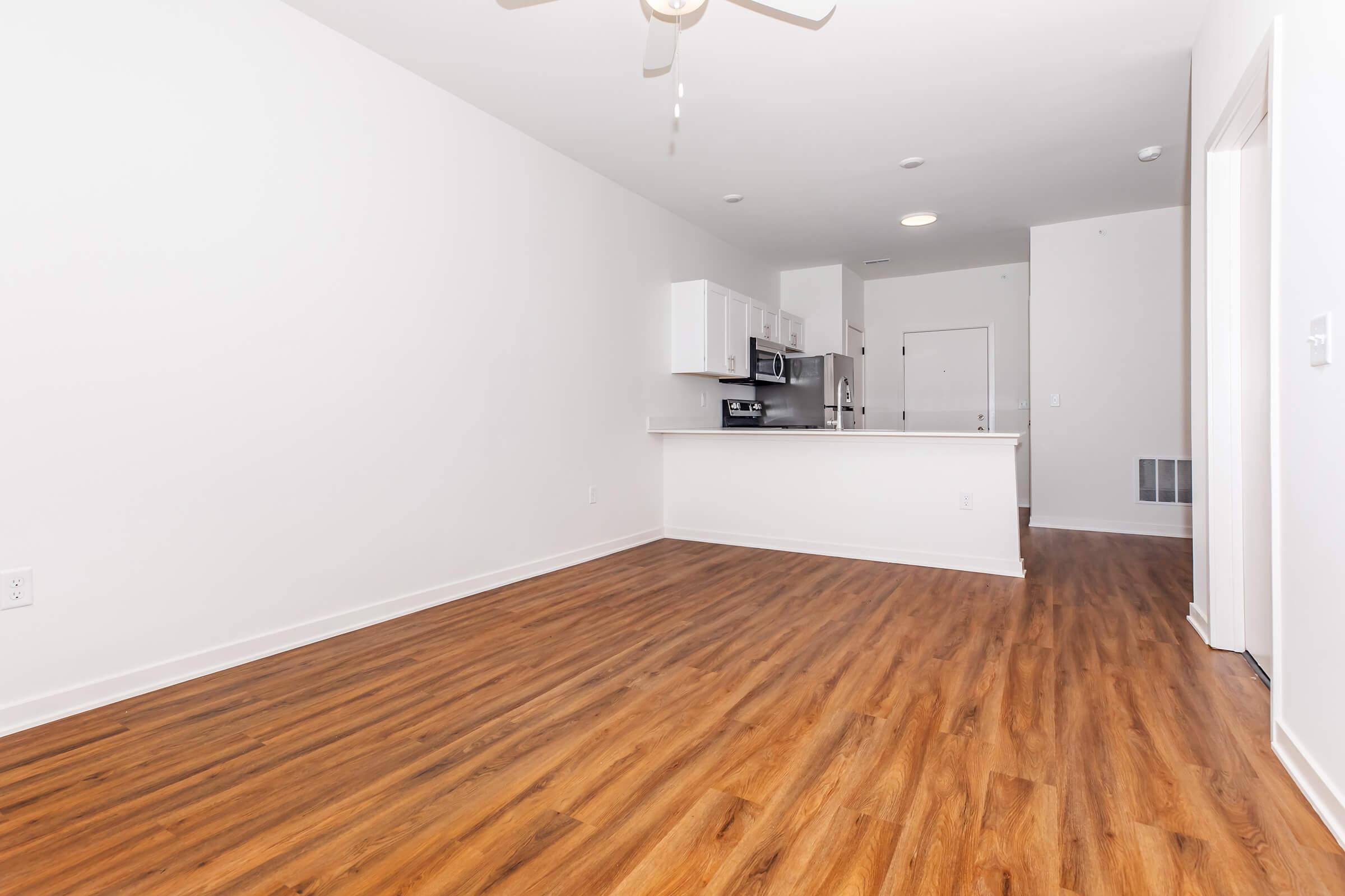 An empty modern living space featuring light-colored walls, hardwood flooring, and a ceiling fan. The open kitchen area includes white cabinets and appliances. Natural light illuminates the room, creating a spacious and inviting atmosphere.