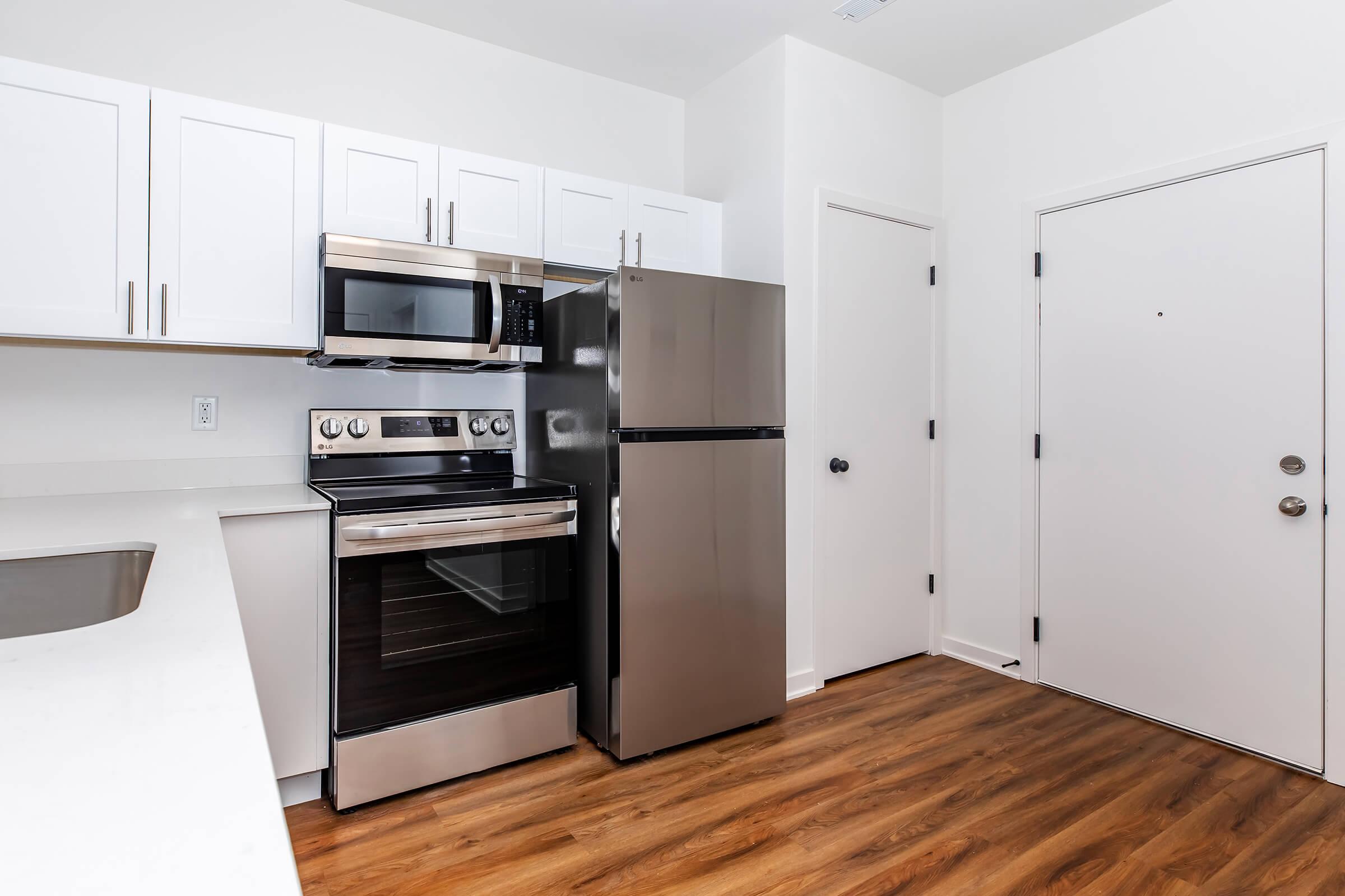 Modern kitchen featuring stainless steel appliances, including a microwave, oven, and refrigerator. The cabinetry is white, and the countertop is a light color. The floor is wooden, and there is a door leading outside, with a simple interior design.