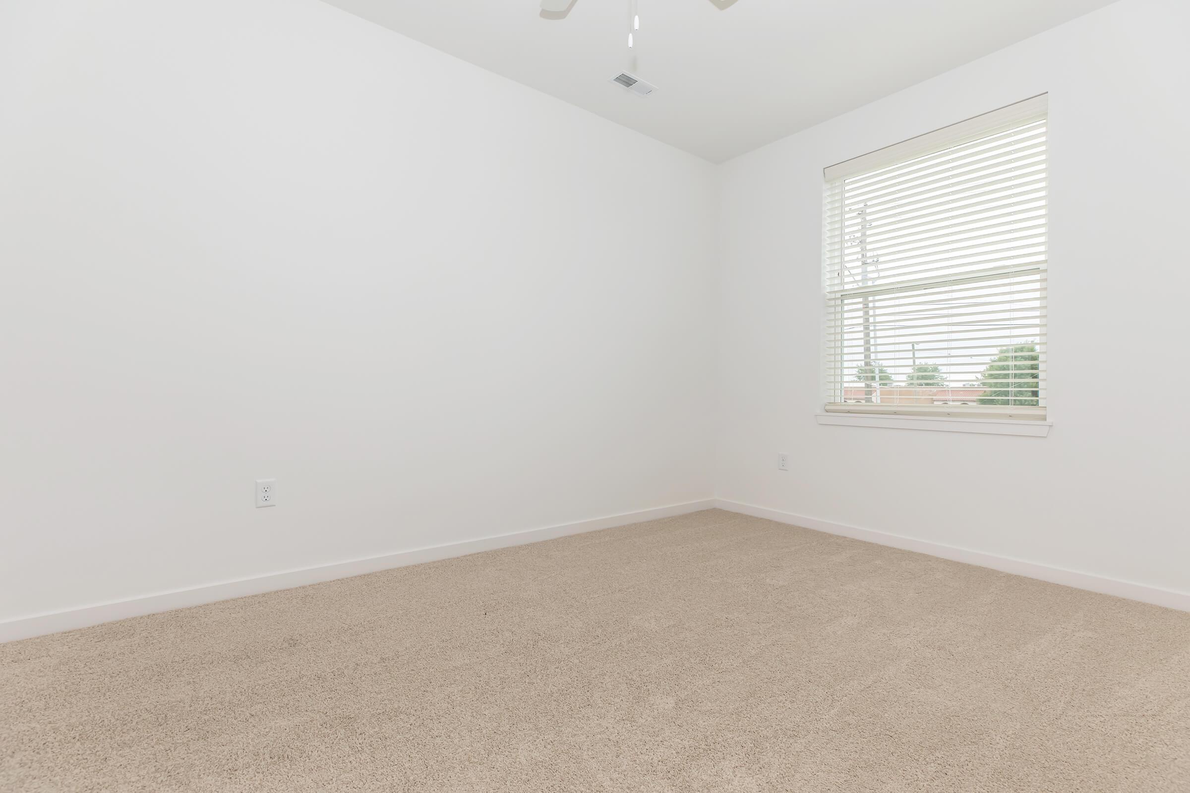 A vacant room with light-colored carpet and white walls. There is a window with blinds on the right side, letting in natural light. The ceiling features a ceiling fan, and the space appears clean and minimalist, suitable for various uses.