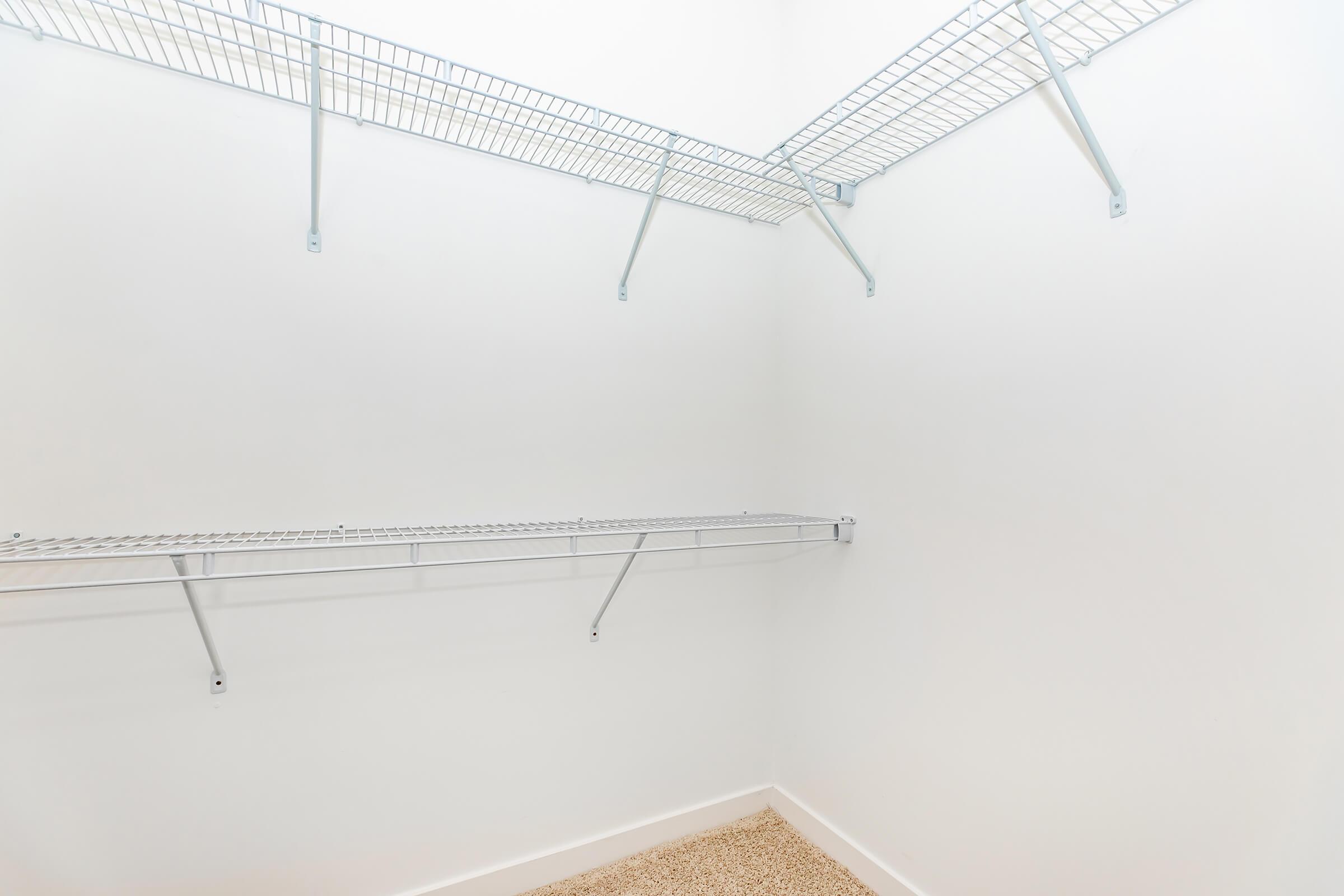 A clean, empty closet with white walls and two wire shelving units hung on the upper section. The floor is carpeted, and there is an overall bright and spacious appearance.