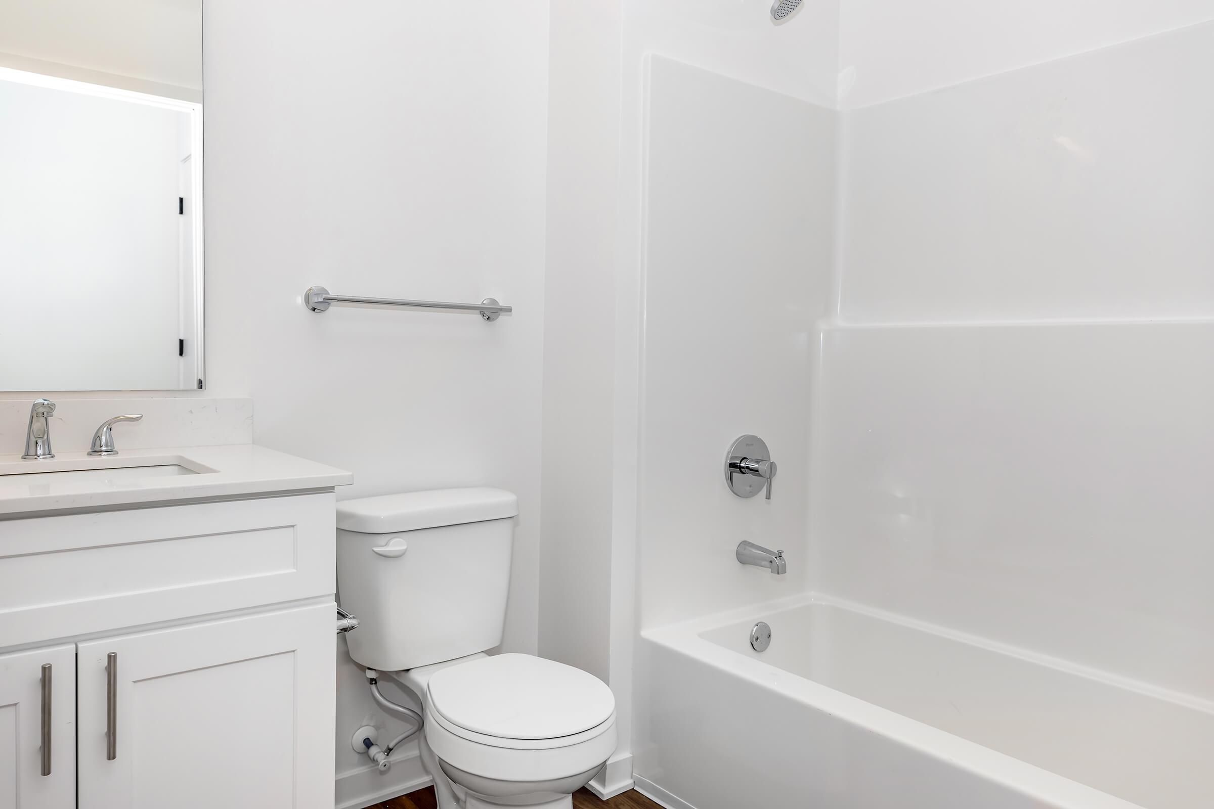 A clean and modern bathroom featuring a white bathtub with a shower, a white toilet, and a sink with a mirror above. The walls and fixtures are all in light tones, creating a bright and spacious feel. The flooring is wooden, adding warmth to the space.