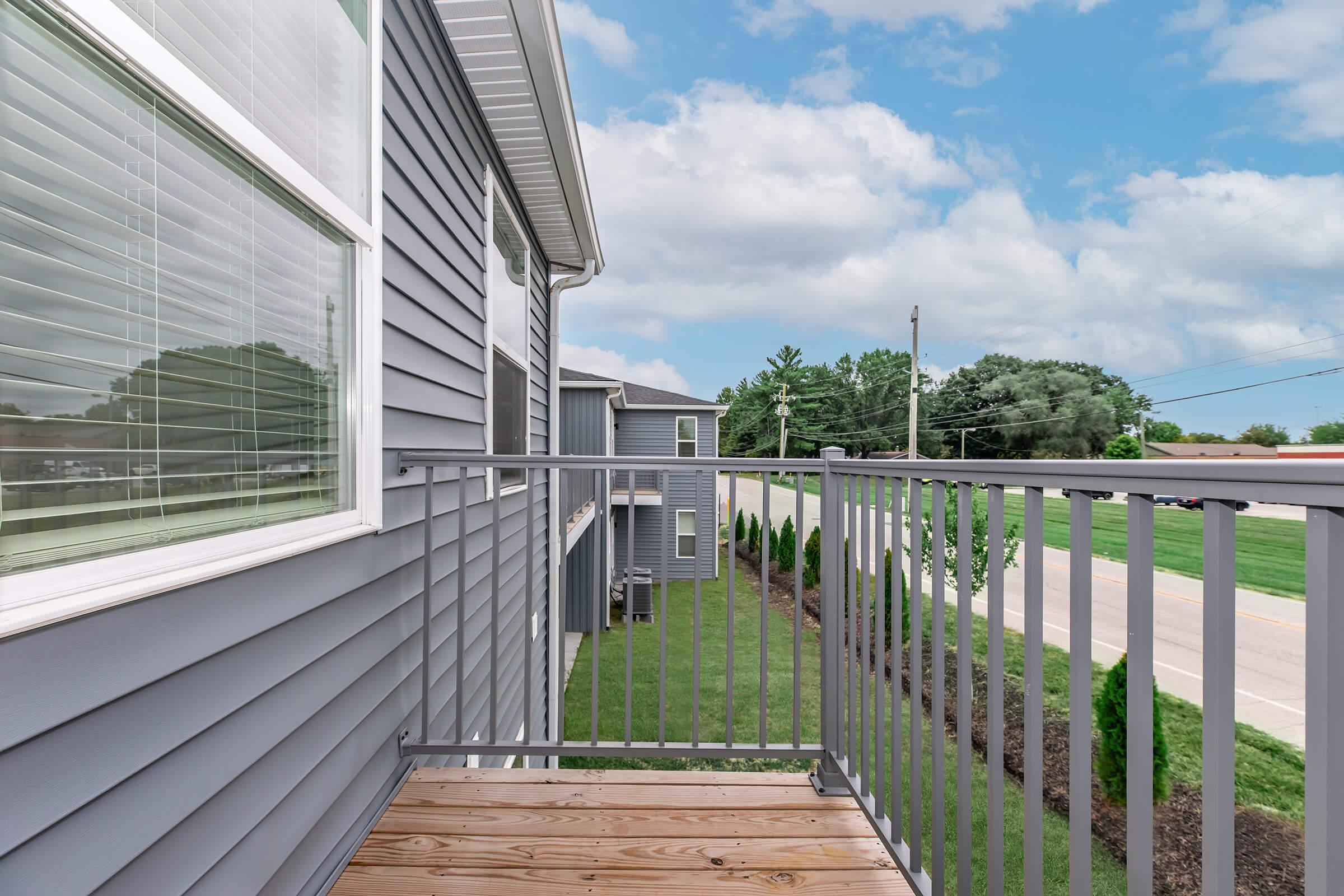 A view from a balcony of a gray-sided building, featuring wooden flooring and a metal railing. In the background, trees and a grassy area are visible, along with a street and power lines under a partly cloudy sky.