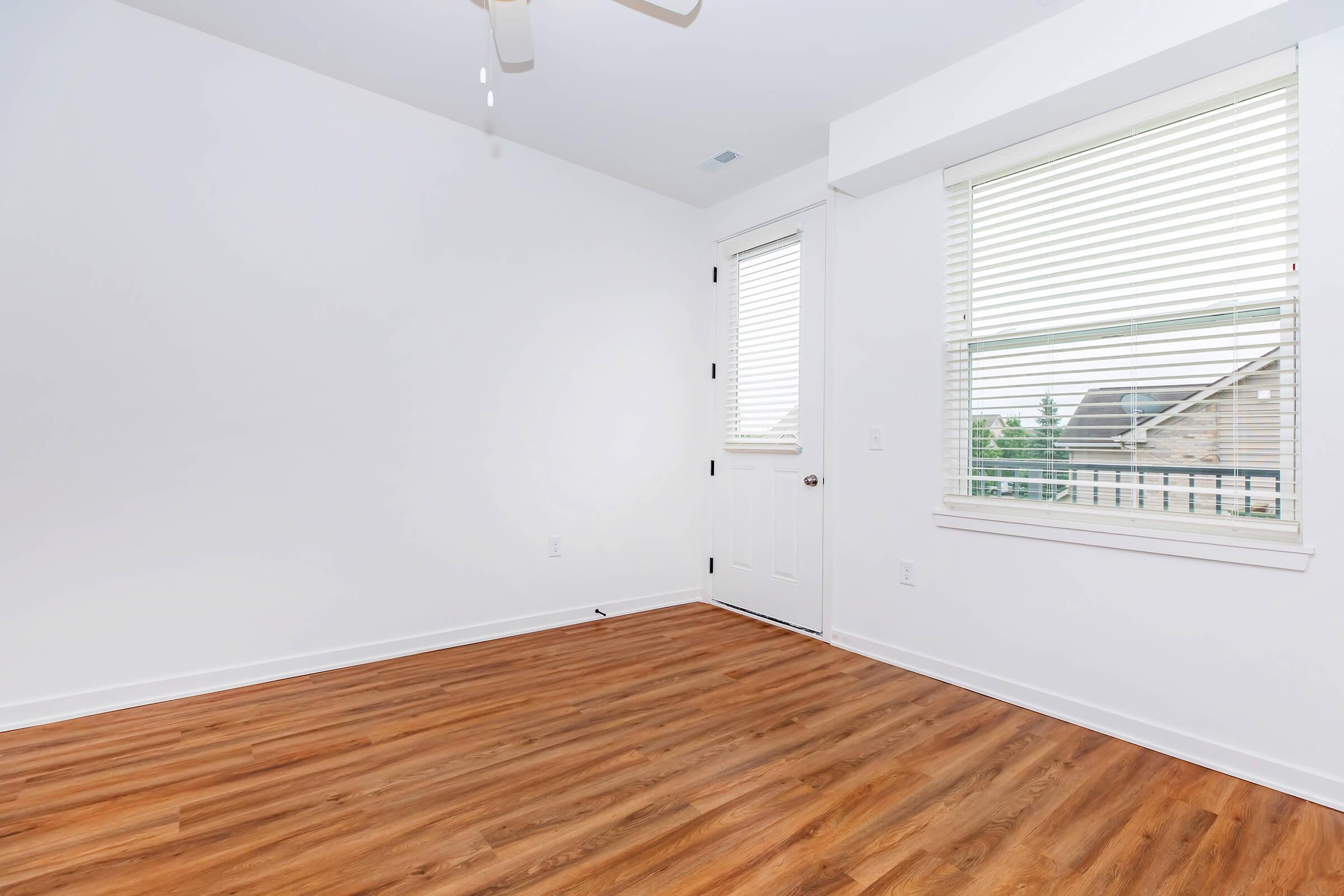 A bright, empty room featuring light-colored walls, a ceiling fan, and hardwood flooring. There is a window with blinds allowing natural light, and a closed door leading outside. The space is uncluttered, offering a fresh and open atmosphere.