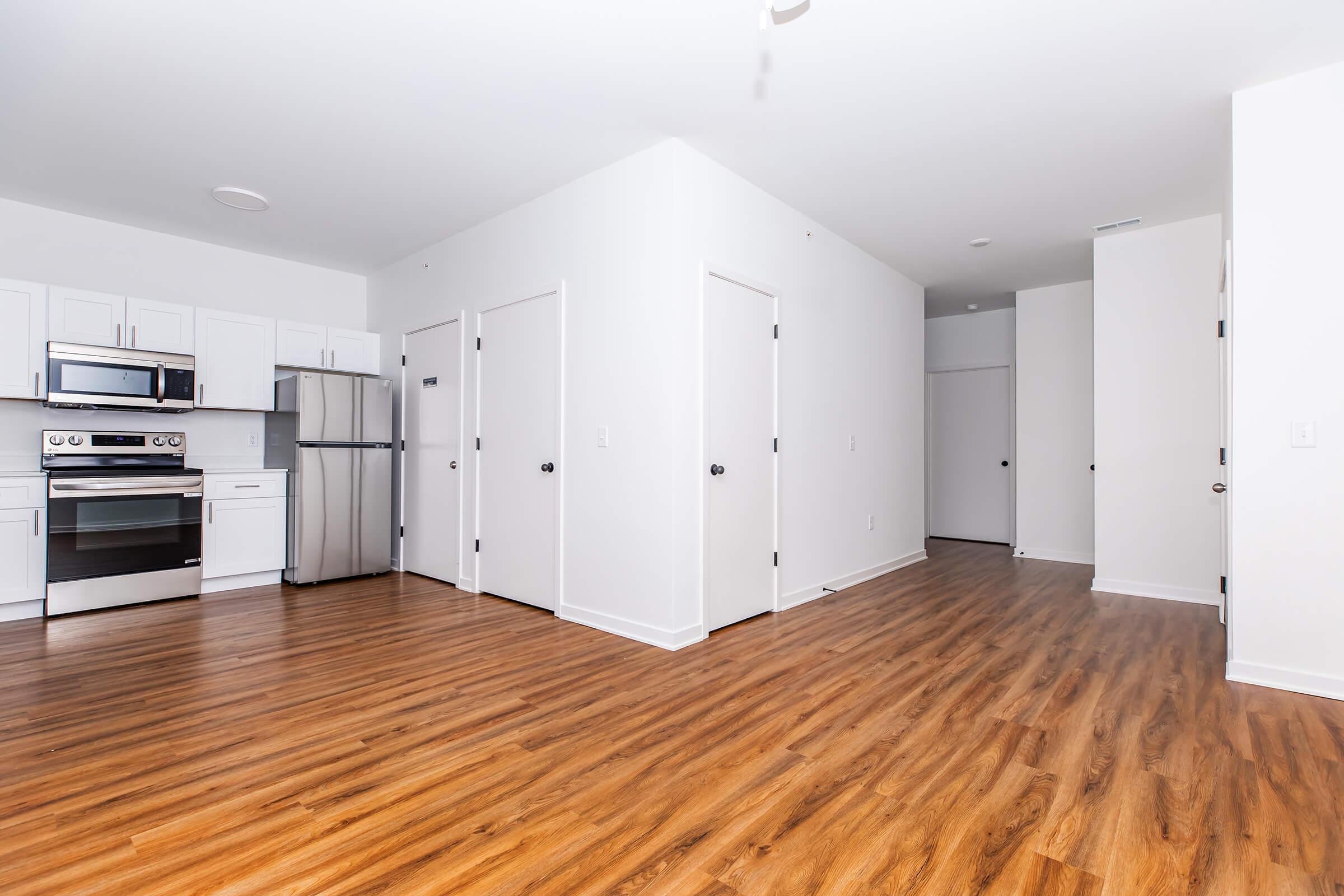 A modern, empty apartment featuring a kitchen with stainless steel appliances, white cabinetry, and a spacious living area with wood-style flooring. Doors lead to adjacent rooms, providing a bright and open atmosphere with neutral-colored walls.