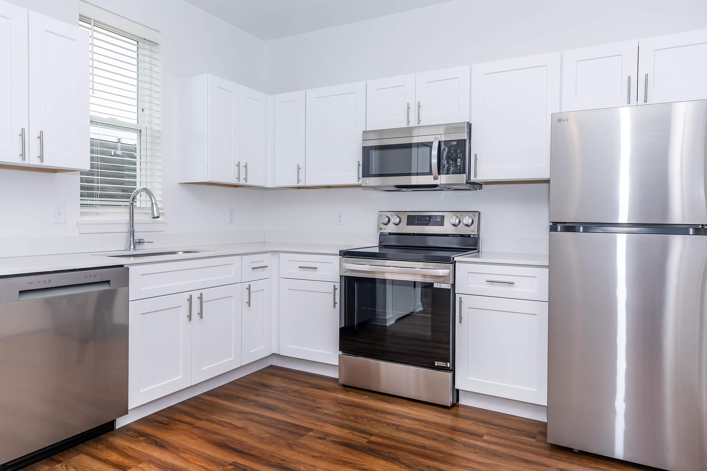 Modern kitchen featuring white cabinetry, stainless steel appliances, including a microwave, oven, and refrigerator. The countertop is light-colored, with a sink under a window. The floor is a warm wood finish, creating a bright and inviting space. Natural light enhances the clean, contemporary design.
