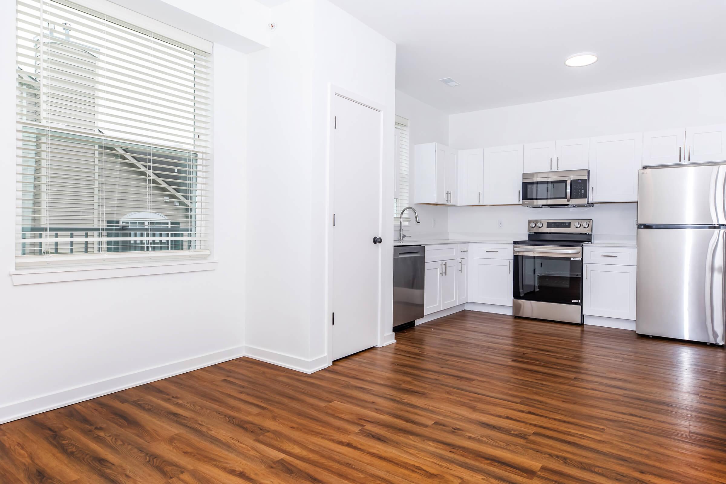 Modern kitchen featuring stainless steel appliances, including a refrigerator, oven, and dishwasher. The space has white cabinetry, a large window with blinds, and warm wood-style flooring. The layout is open, creating a bright and inviting atmosphere.